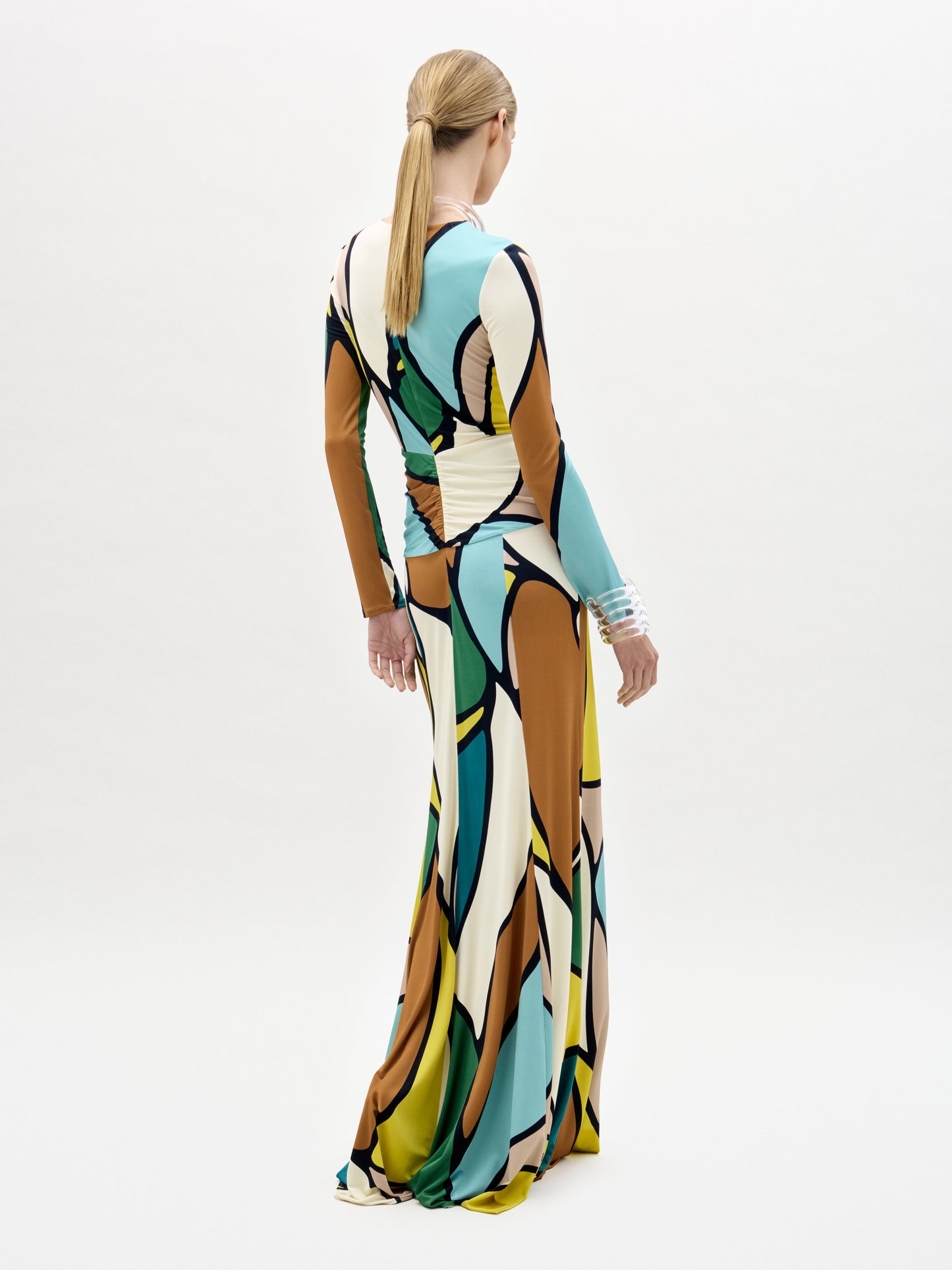Una mujer con el pelo rubio recogido en una coleta baja lleva el vestido Lucian, un maxi largo y entallado con un colorido estampado abstracto confeccionado en tela italiana. Está de pie sobre un fondo blanco liso, de espaldas a la cámara.
