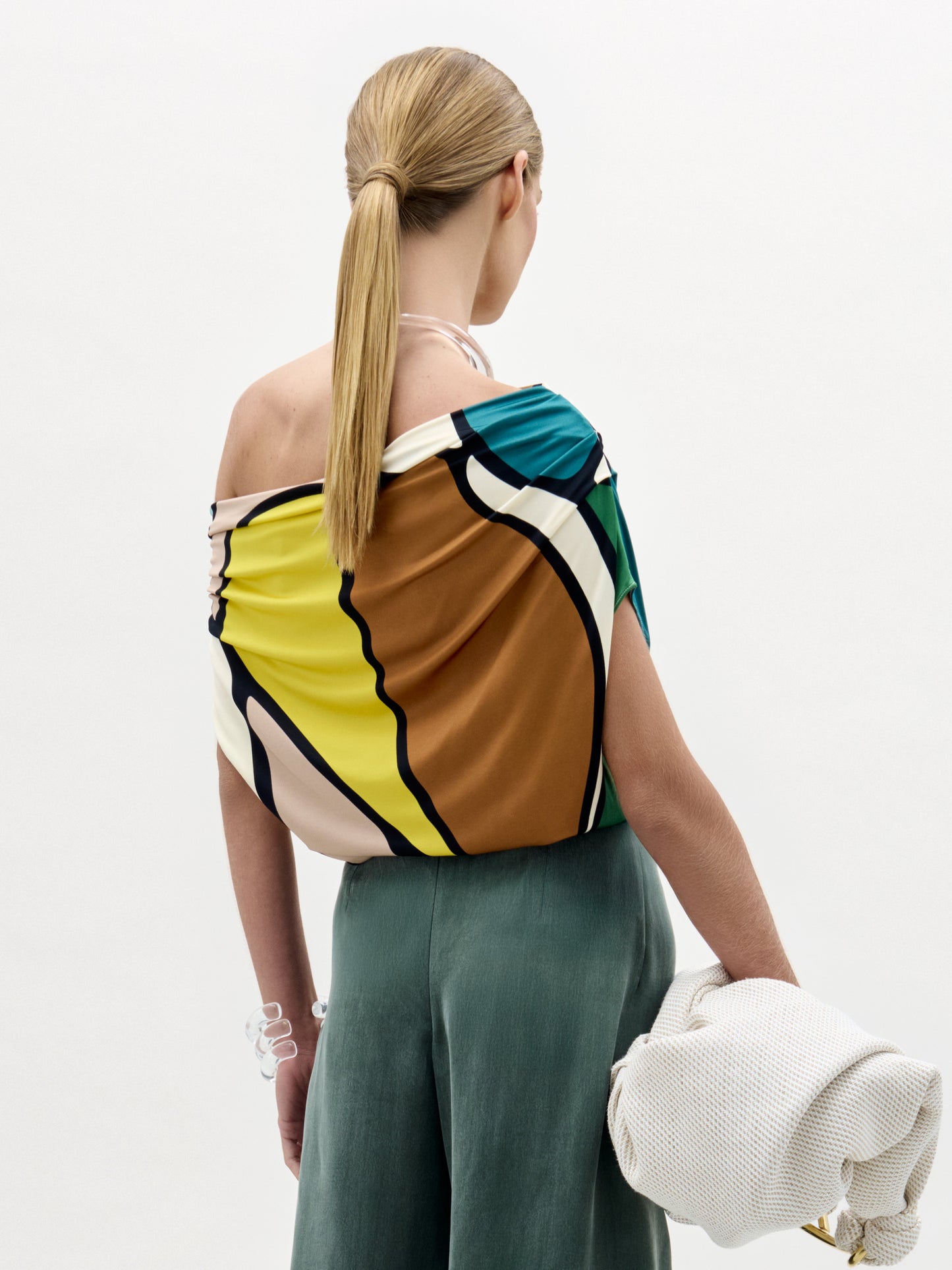 Una persona con el pelo largo y rubio recogido en una coleta baja mira hacia otro lado, llevando la Blusa Indiana -un top asimétrico geométrico y colorido- con pantalones verde salvia y un bolso blanco texturizado para un look effortless hecho en Colombia.