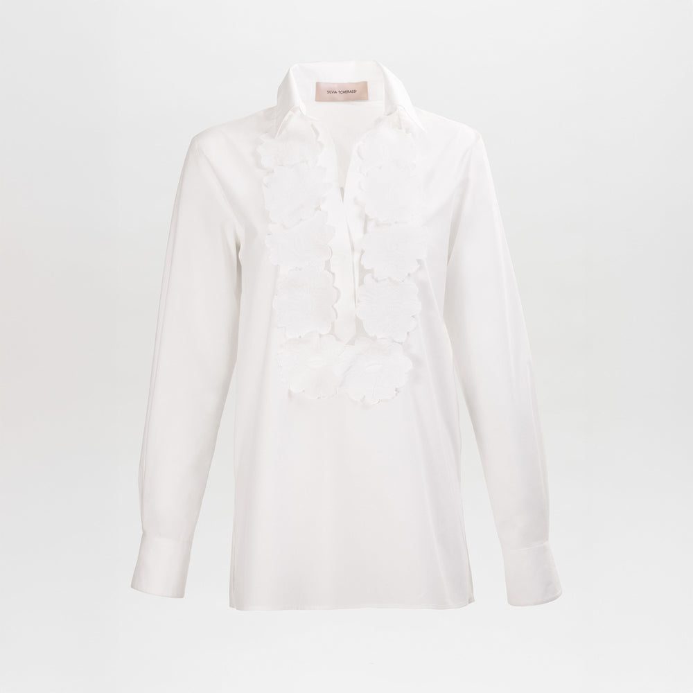 La blusa Karole es una camisa blanca de manga larga que presenta un cuello puntiagudo y llamativos detalles florales aplicados a lo largo de la tapeta delantera, sobre un fondo blanco liso.