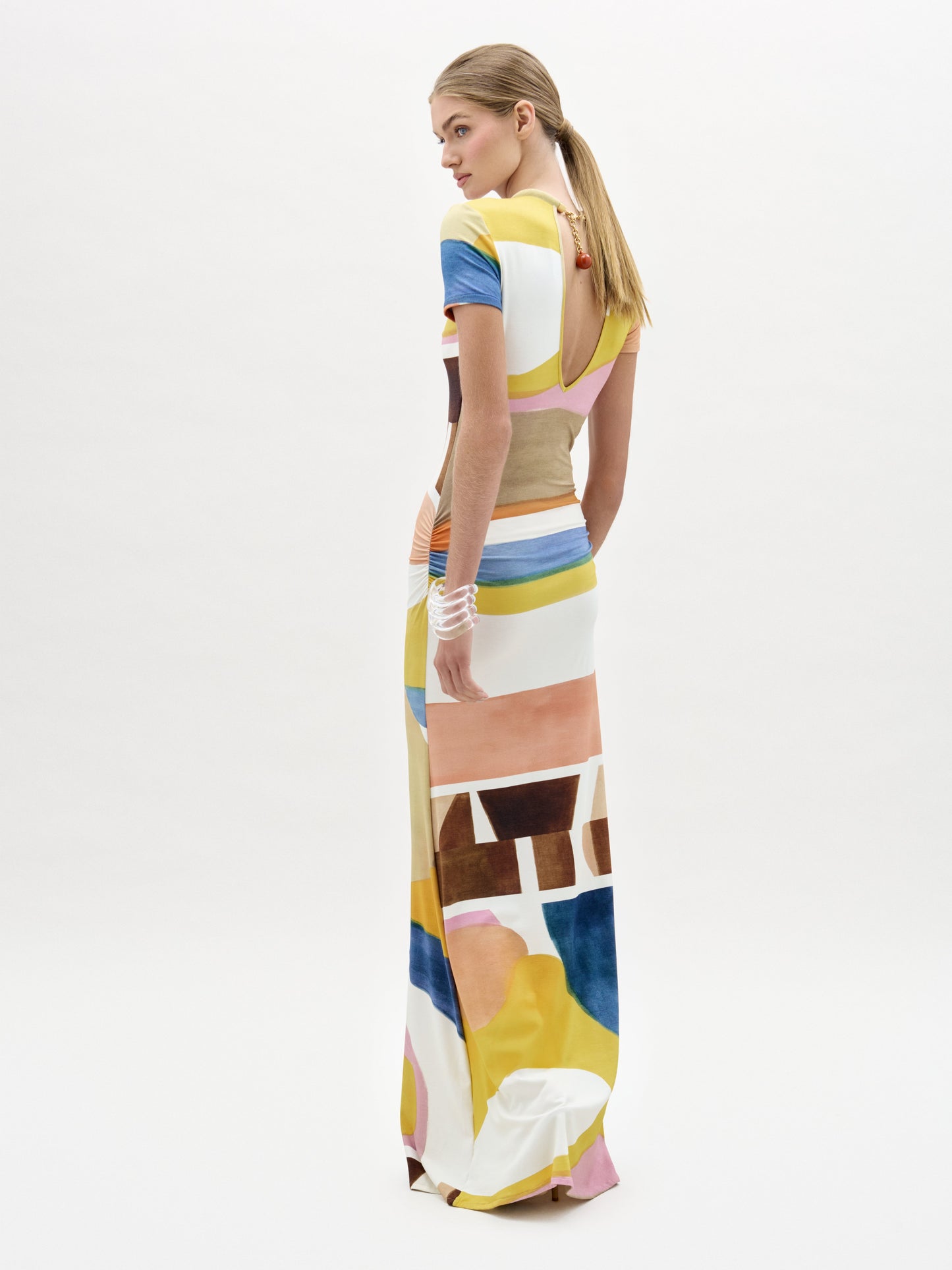 Una mujer con el pelo largo y rubio recogido en una coleta baja lleva el vestido Ikia, un maxi largo hasta el suelo con mangas cortas y un colorido estampado abstracto, mientras mira por encima del hombro sobre un fondo blanco liso.