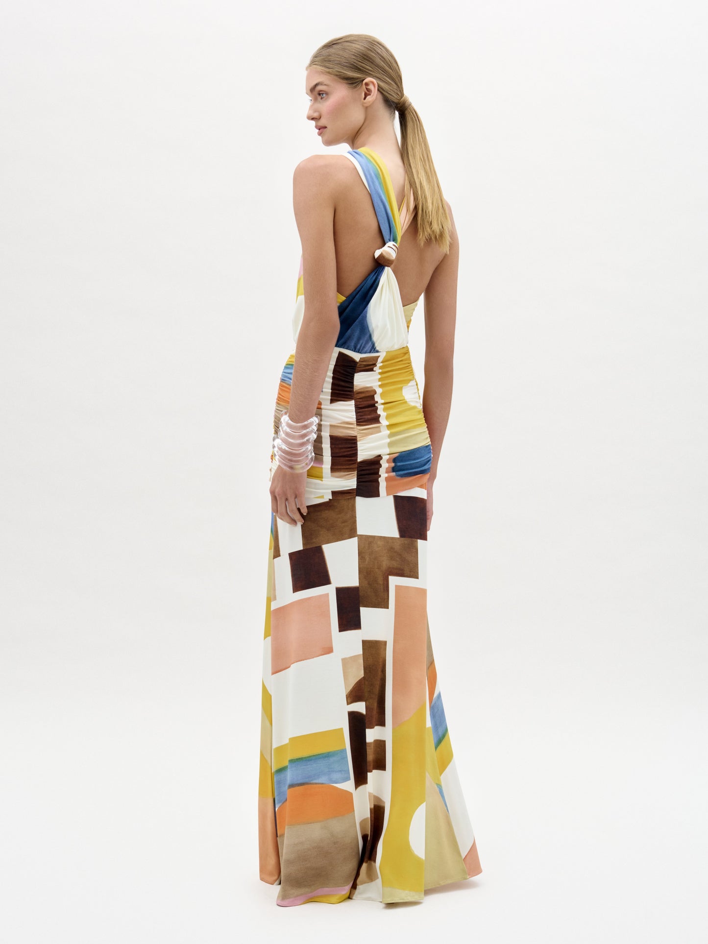 Una mujer de piel clara y pelo liso recogido lleva el vestido Jedda, un maxi ajustado con un estampado geométrico amarillo, marrón, naranja, azul y blanco. Está de lado y lleva varias pulseras en la muñeca izquierda.