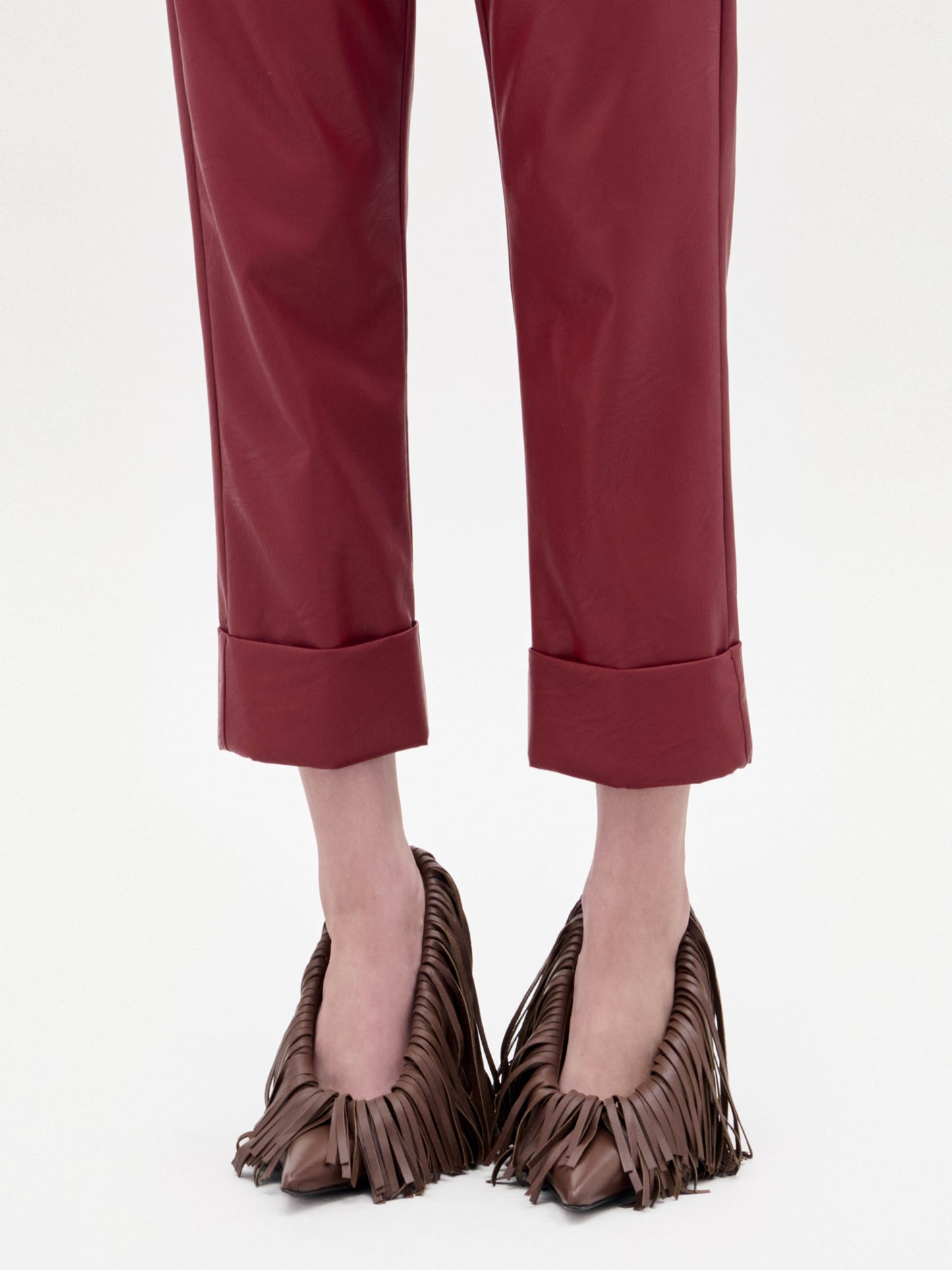 Una persona con pantalones rojos arremangados lleva tacones Amelia -zapatos marrones de tacón alto con llamativos flecos de cuero- mientras permanece de pie sobre un fondo blanco liso.