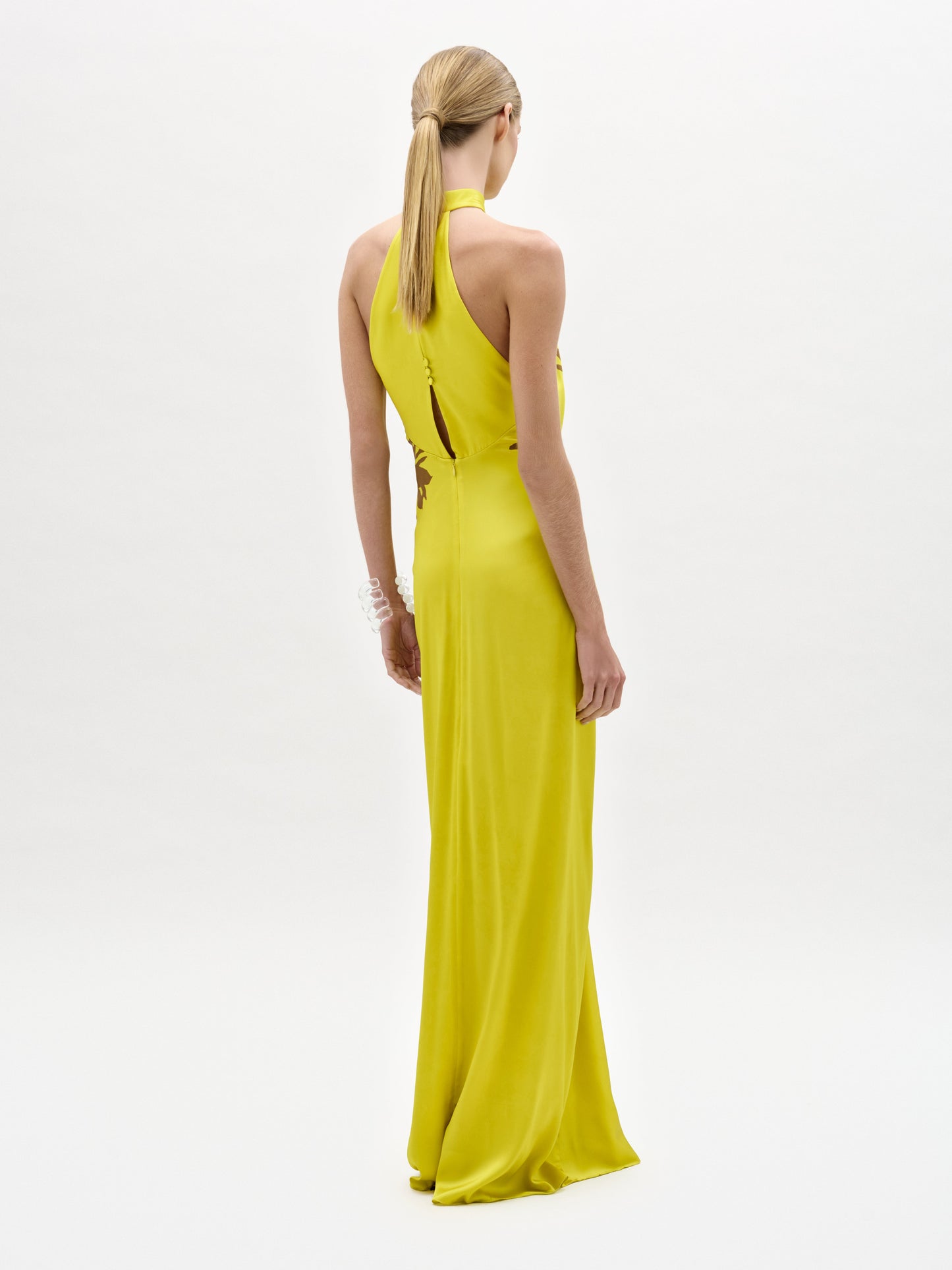 Una persona con el pelo largo y rubio recogido en una coleta lleva el Vestido Koori, un maxi de seda amarilla brillante con escote halter, largo hasta el suelo, de pie sobre un fondo blanco liso y de espaldas a la cámara.