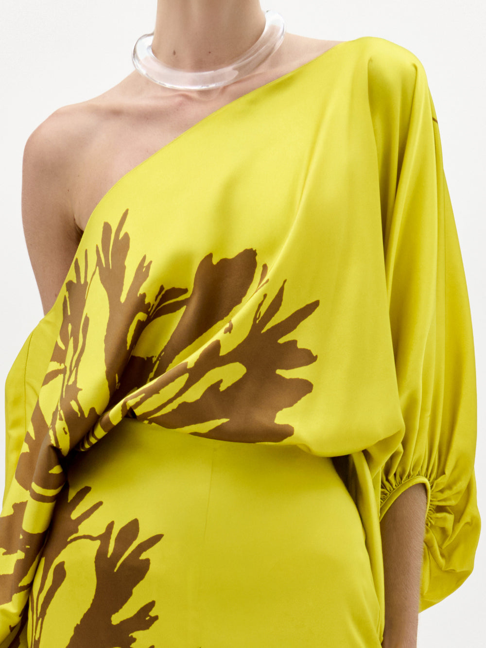 Una persona lleva la blusa Bellagio, un top amarillo de un solo hombro con motivos abstractos marrones en tela italiana, que presenta un dobladillo fruncido en las mangas y se combina con un collar de trozos transparentes sobre un fondo blanco.