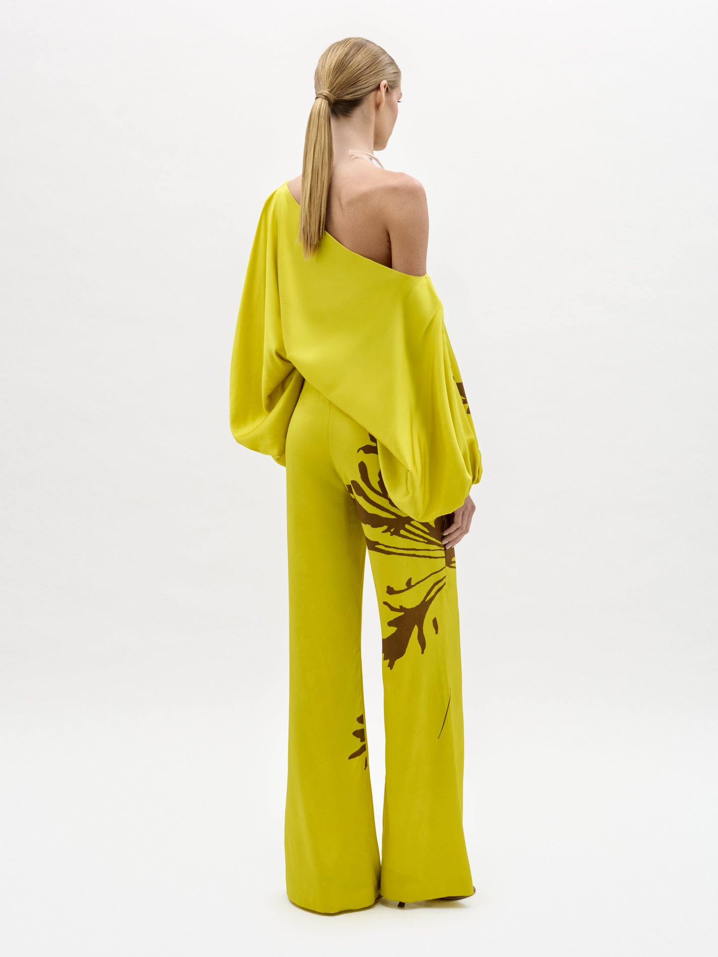Una persona con el pelo rubio y liso recogido en una coleta baja lleva un top suelto amarillo brillante de un solo hombro con un pantalón Andie a juego con motivos florales oscuros y botas anchas, de pie sobre un fondo blanco liso.