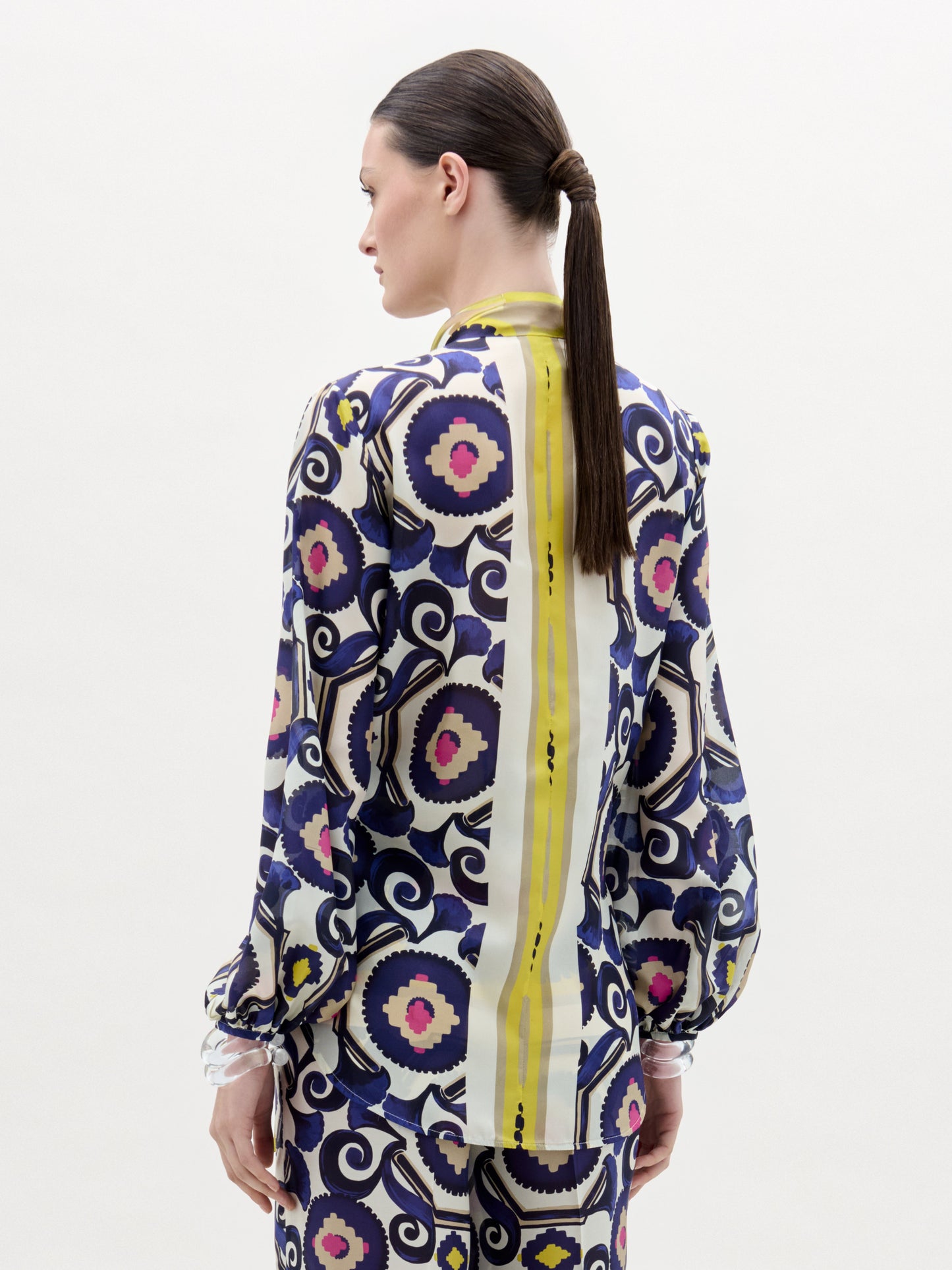 Una mujer con el pelo castaño liso recogido en una coleta baja mira de frente a la cámara, llevando la Blusa Miel -una blusa de manga larga hecha en Colombia con diseños en morado, amarillo, rosa y azul marino- sobre un fondo liso.