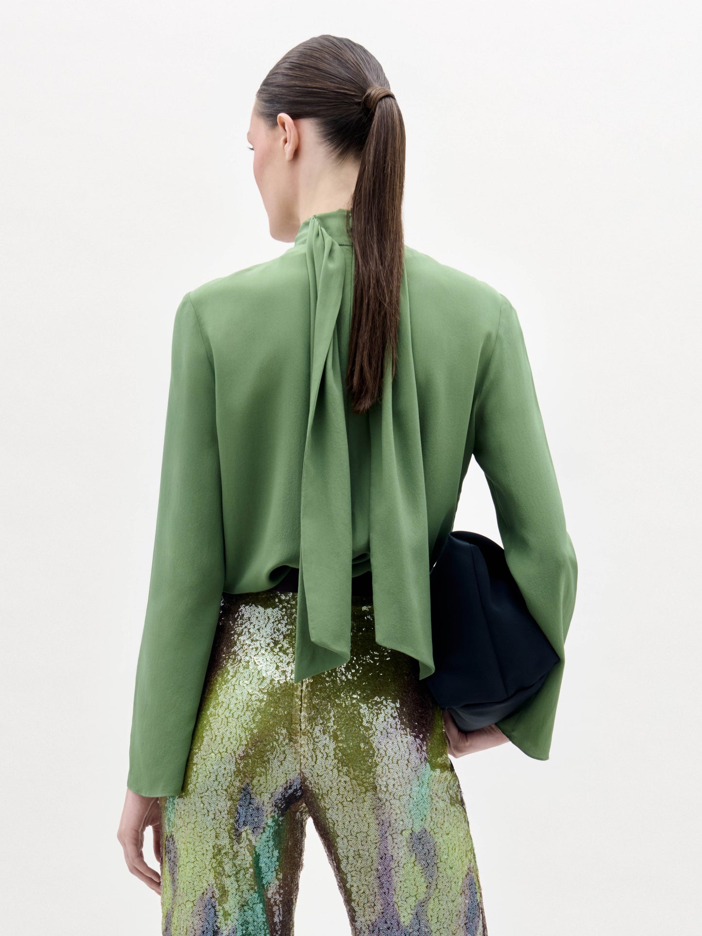 Se ve de espaldas a una persona con el pelo largo y oscuro recogido en una coleta baja, que lleva la blusa Kelsey verde con un lazo en el cuello, pantalones de lentejuelas multicolores y un bolso de mano negro.