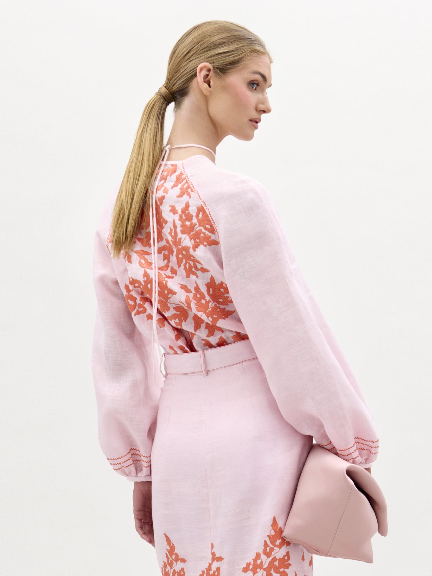 Una mujer con el pelo rubio recogido en una coleta baja lleva la blusa Molveno, un top de lino rosa claro con motivos florales naranjas y mangas largas. Lleva un bolso de mano rosa pálido y está de pie sobre un fondo liso y claro, mirando ligeramente hacia otro lado.