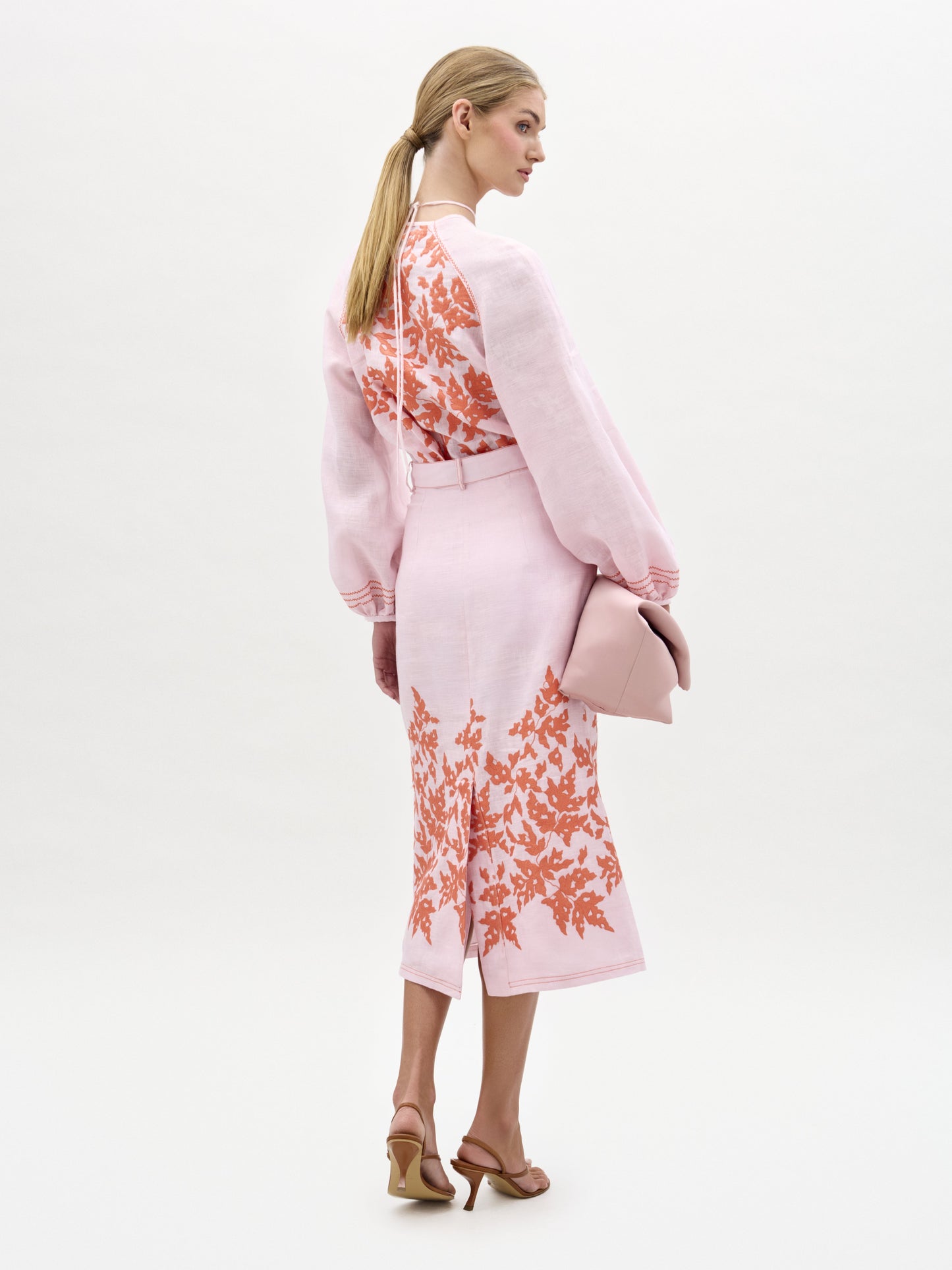 Una mujer mira de frente con la falda Layan, una falda midi rosa claro con estampado floral naranja. La lleva con mangas largas, un bolso de mano rosa a juego, sandalias de tacón color canela y el pelo rubio recogido en una coleta baja.