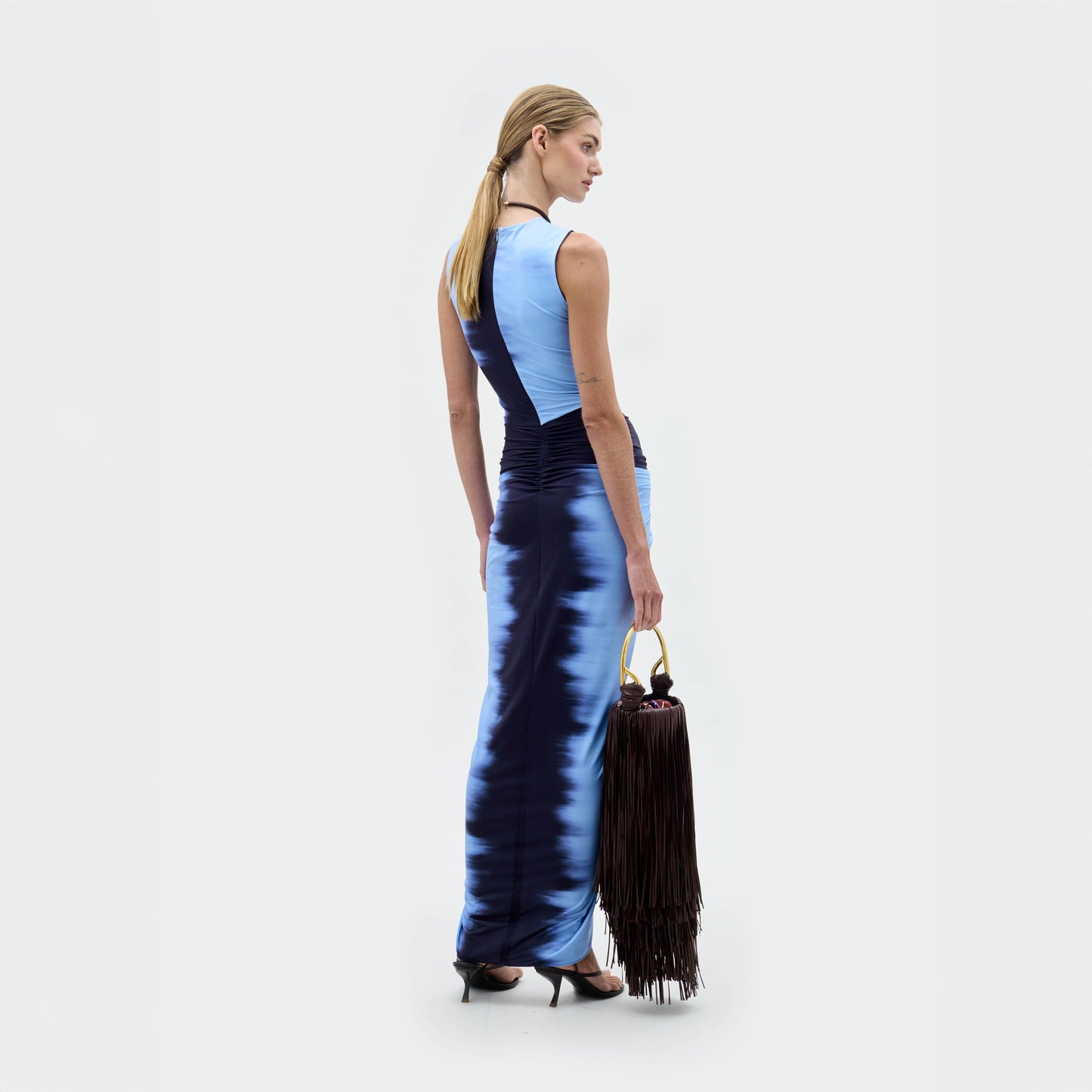 Una mujer de espaldas sobre un fondo blanco liso, con el vestido Leni sin mangas hasta el suelo en degradado azul. Lleva un gran bolso marrón con flecos, el pelo rubio recogido en una coleta baja y tacones negros.