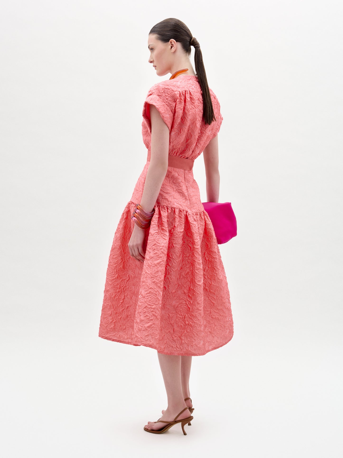 Una mujer de perfil luce el vestido Metaponto, un midi coral con textura, cintura entallada y falda acampanada. Lleva un bolso de mano rosa brillante, sandalias de tacón marrones y el pelo recogido en una coleta baja.