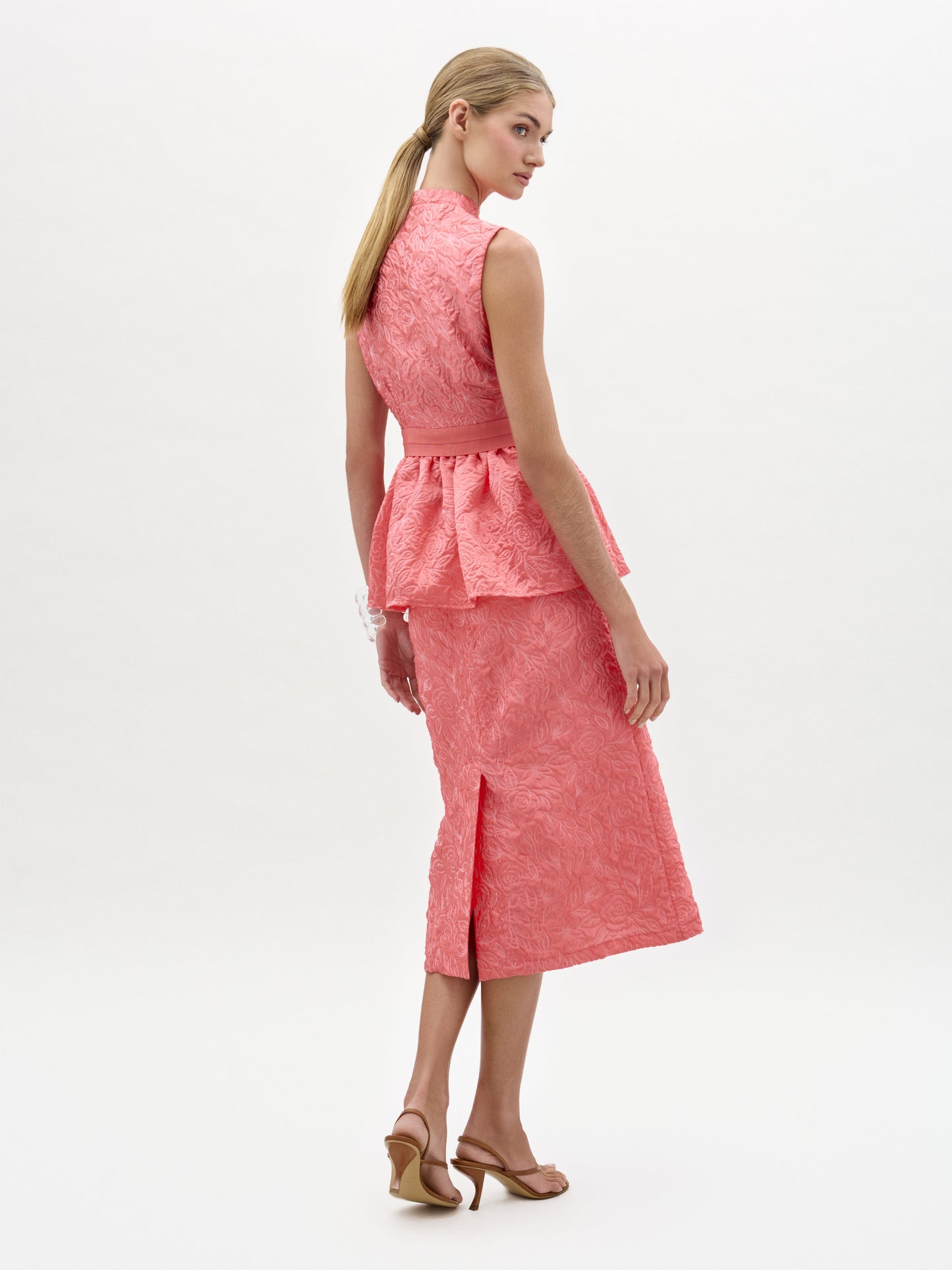 Una mujer mira por encima del hombro, con un top coral texturizado sin mangas y la falda Atira -una falda midi a juego con abertura trasera en tejido italiano, confeccionada en Colombia- combinada con sandalias de tacón beige sobre un fondo blanco liso.