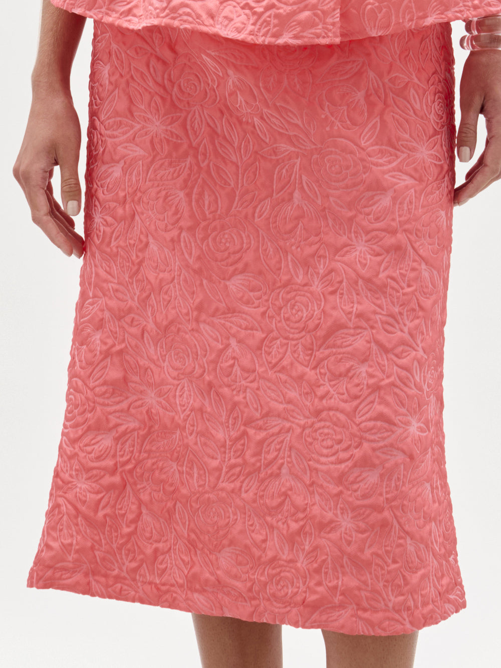 Una persona lleva la falda Atira en rosa coral, una falda midi hasta la rodilla con un estampado floral en relieve confeccionado en tejido italiano. Sus manos están a los lados sobre un fondo blanco liso.