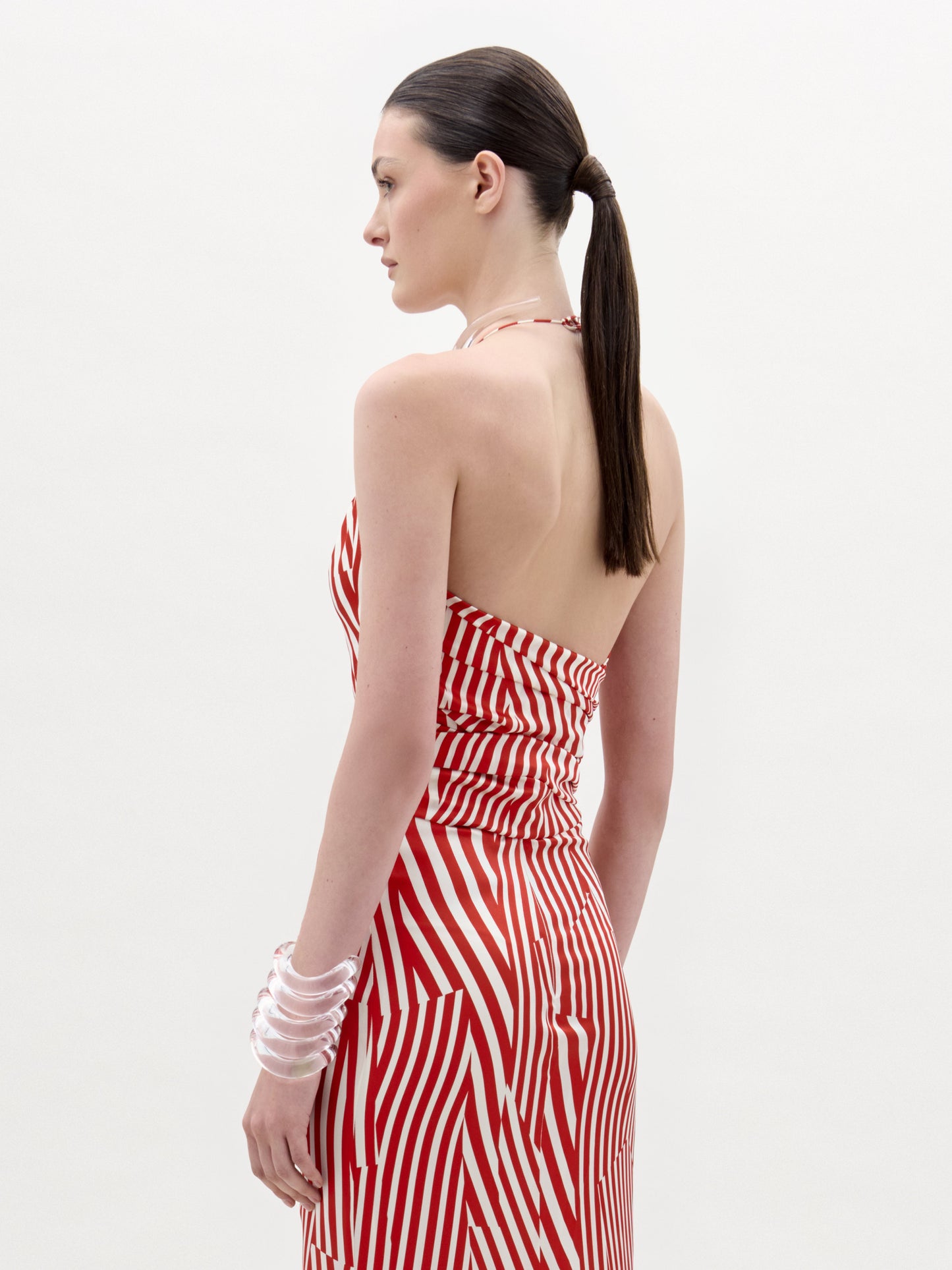 Una mujer con el pelo largo y oscuro recogido en una coleta baja aparece de perfil, con la blusa Carmela -una blusa de tirantes a rayas rojas y blancas con la espalda abierta- y brazaletes transparentes en las muñecas, sobre un fondo claro y liso.