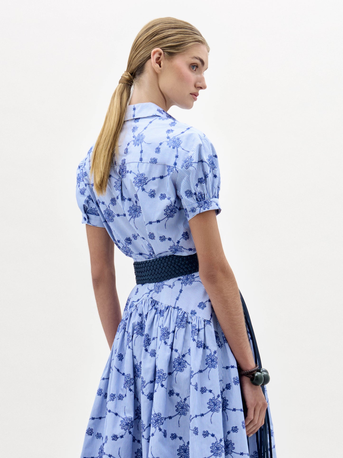 Una mujer con el pelo castaño claro y liso recogido en una coleta baja lleva la blusa Ileana -de flores azules y blancas con mangas cortas y un cinturón tejido oscuro- de pie sobre un fondo liso, mirando a su derecha.