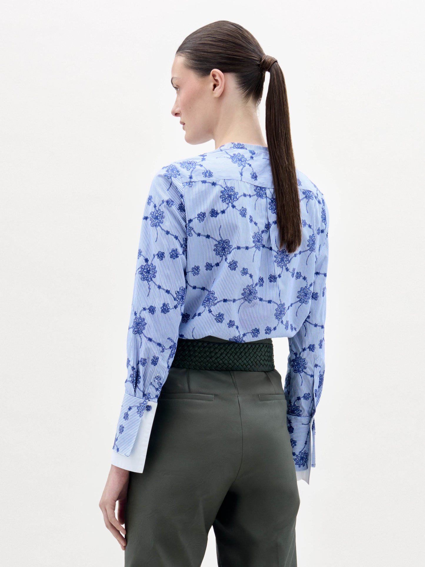 Una mujer con el pelo castaño liso recogido en una coleta baja aparece de espaldas, con la blusa Cubillos -una camisa de manga larga de flores azules y blancas- metida dentro de un pantalón verde oliva de cintura alta con un cinturón de textura oscura sobre un fondo blanco liso.