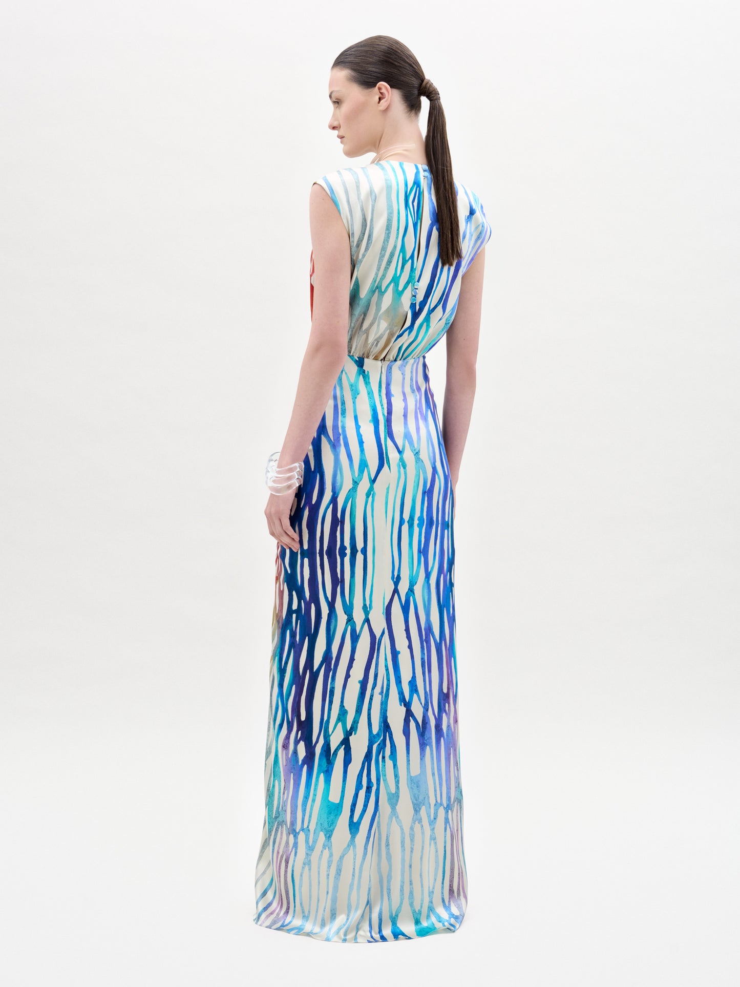 Una persona mira hacia otro lado y lleva el vestido Samaira, un maxi largo con estampado abstracto azul y morado y escote en forma de ojo de cerradura. Lleva el pelo recogido en una coleta baja y una pulsera blanca. El fondo es blanco.