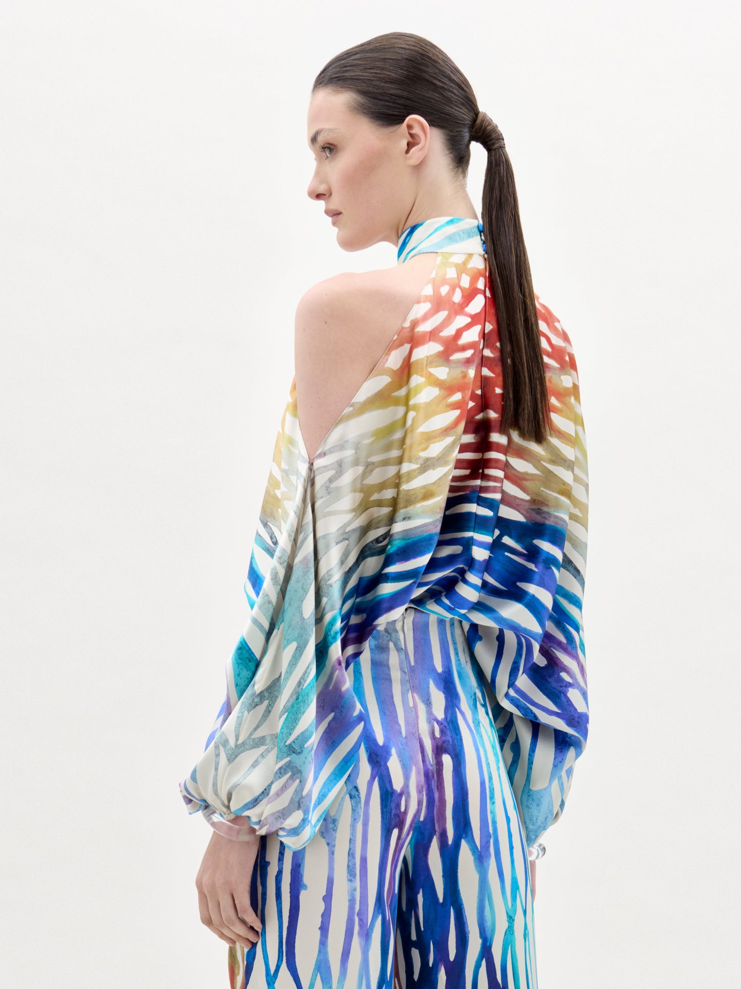 Una persona con el pelo largo y oscuro recogido en una coleta baja se ve de perfil y de espaldas, llevando la blusa Safira, una colorida blusa de cuello alto con un estampado abstracto de rayas y pinceladas azules, rojas, amarillas y blancas.