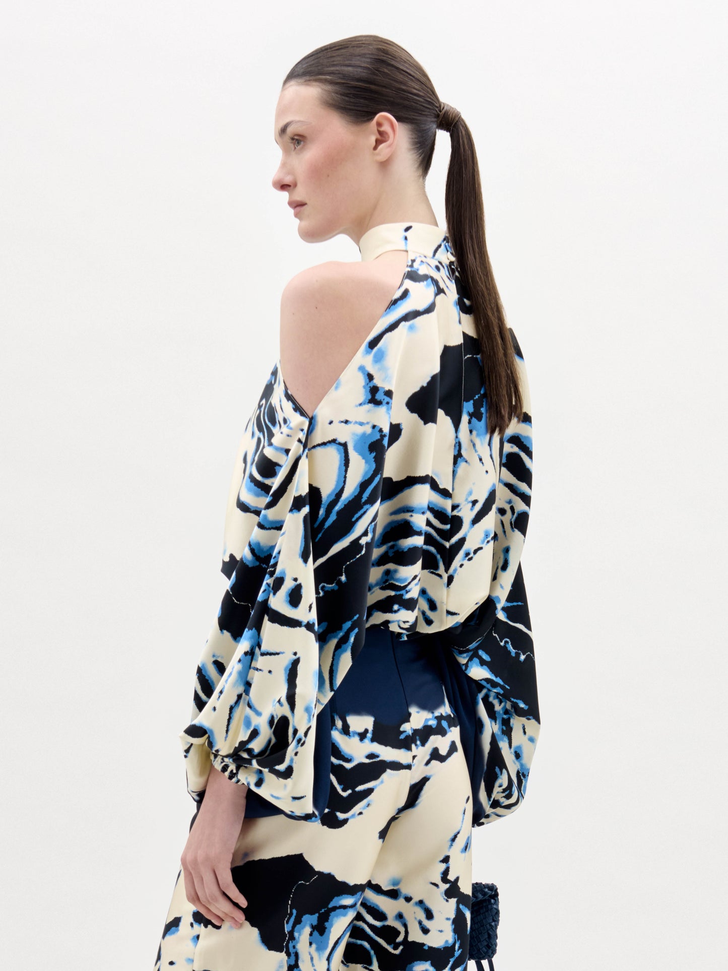 Una persona con el pelo largo y castaño recogido en una coleta baja lleva la blusa Safira, un top de cuello halter con motivos abstractos en azul, negro y crema y un hombro al descubierto. El fondo es blanco liso.