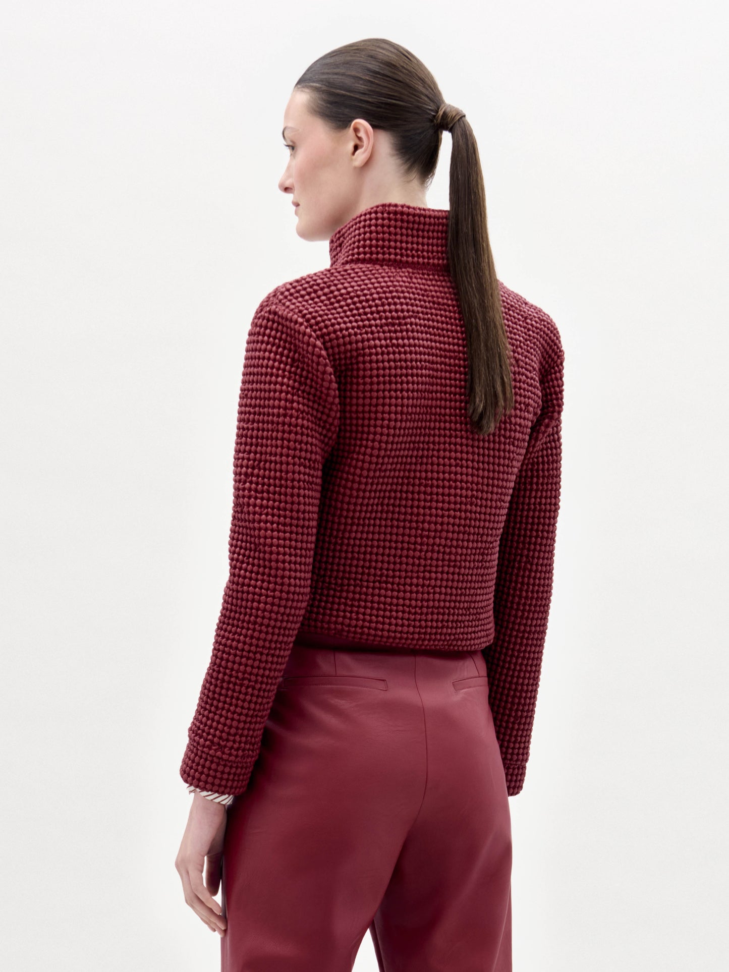 Una persona con el pelo largo y castaño recogido en una coleta baja se ve de espaldas, vistiendo la chaqueta Zalta texturizada de color rojo oscuro y pantalones a juego sobre un fondo blanco liso.