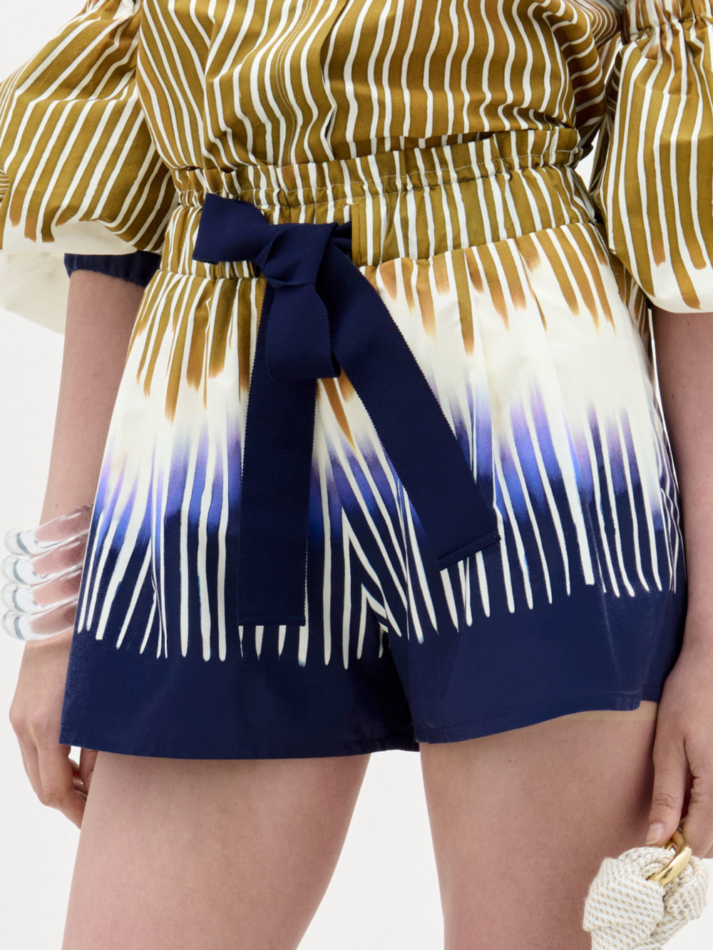 Una persona lleva el pantalón corto Giorgio, un conjunto de cintura alta con rayas azul marino, blancas y marrones, peinado con un cinturón azul oscuro, pulseras de trozos transparentes y un pequeño bolso con textura. Fabricado en Colombia con algodón 100% para mayor comodidad y estilo.