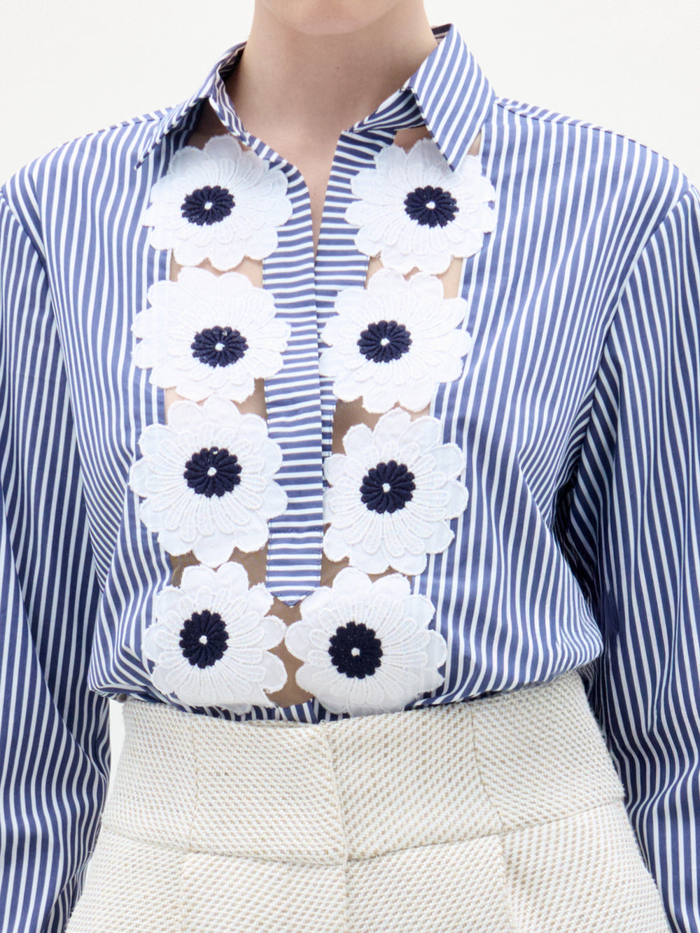 Una persona lleva la blusa Karole, una prenda abotonada de rayas verticales azules y blancas con grandes apliques florales blancos confeccionada en Colombia, combinada con unos pantalones de cintura alta de textura crema; no se muestra su rostro.