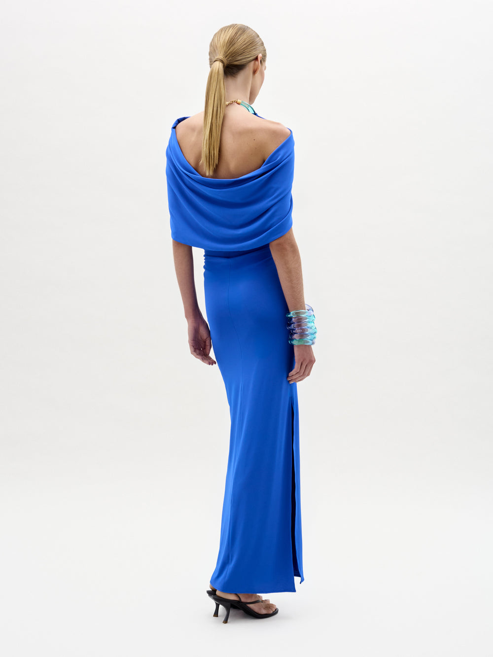 Una persona mira hacia otro lado, lleva el vestido Odele -un maxi azul brillante sin hombros con una abertura lateral- confeccionado en tela italiana. Lleva el pelo rubio liso recogido en una coleta baja, tacones negros y varias pulseras azules en un brazo.