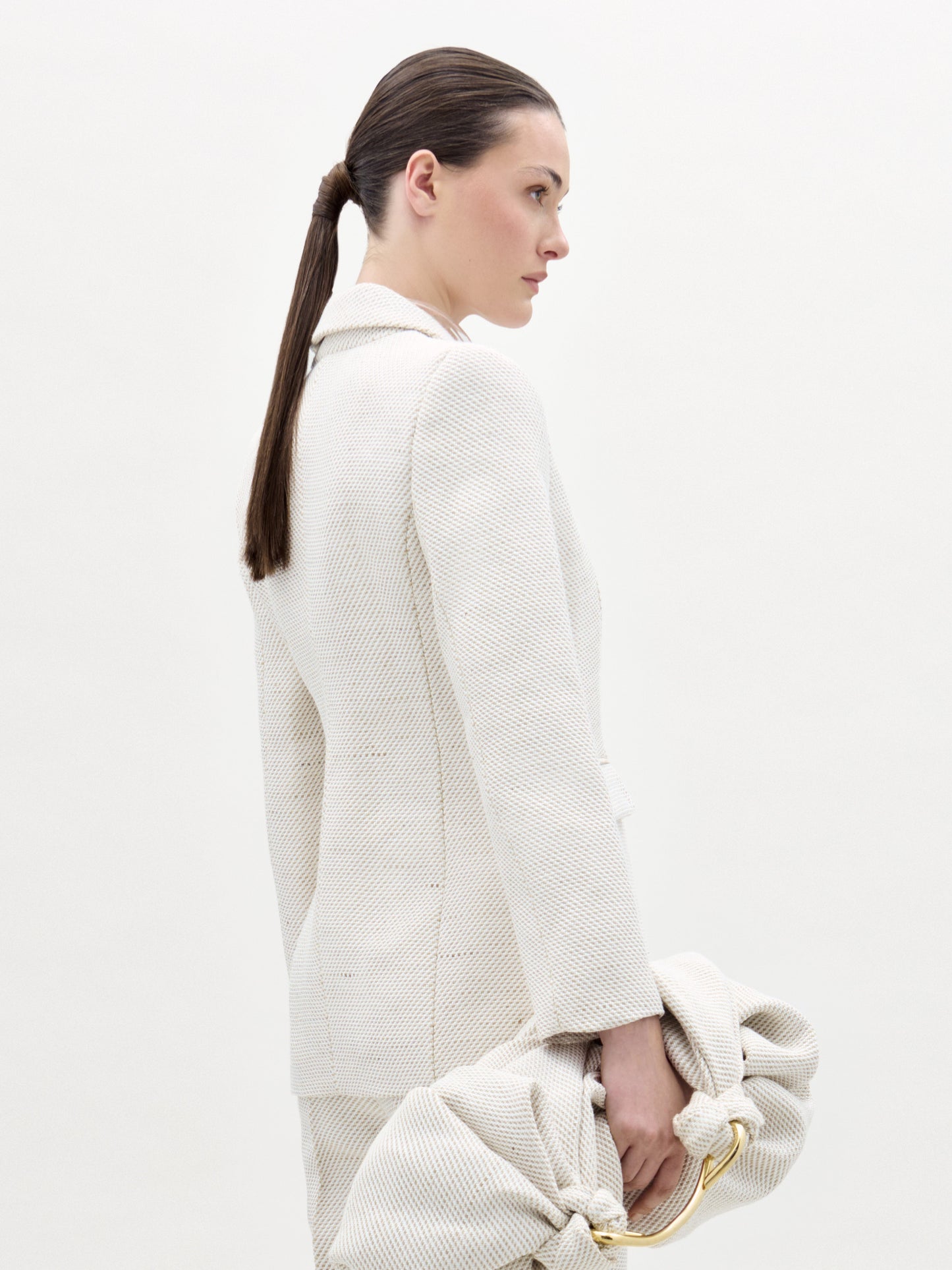 Una mujer con el pelo largo y castaño recogido aparece de perfil, con la chaqueta Rebeca entallada y un bolso de mano con textura a juego, sobre un fondo liso de color claro.