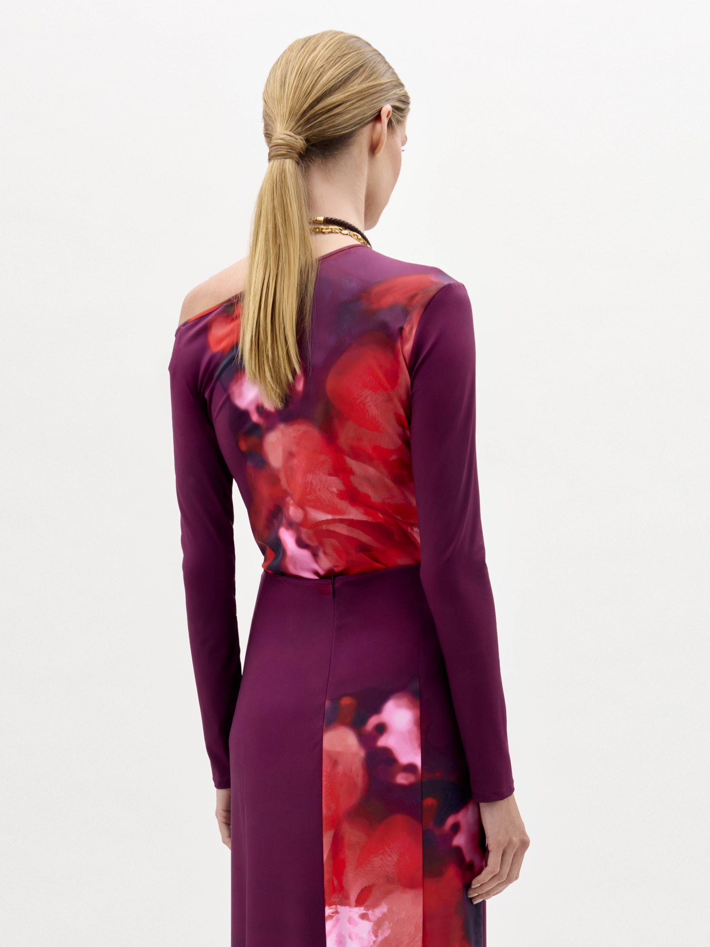 Una persona con el pelo rubio recogido en una coleta baja, de pie y de espaldas, lleva la blusa Sabra, de manga larga y color morado, con un estampado floral abstracto rojo y rosa en la espalda. El fondo es liso y de color claro.