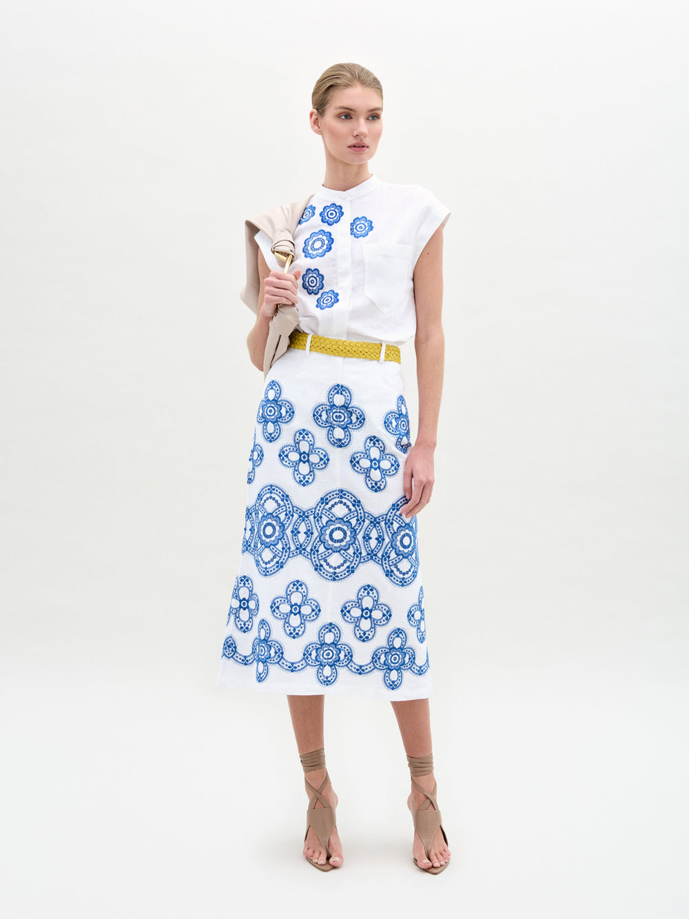 Una persona está parada sobre un fondo blanco con una elegante blusa blanca y una falda midi Falda Layan con estampados florales azules. Sostiene una chaqueta beige doblada sobre su hombro izquierdo, mostrando la moda de Hecho en Colombia, completada con un cinturón amarillo y sandalias de tiras.