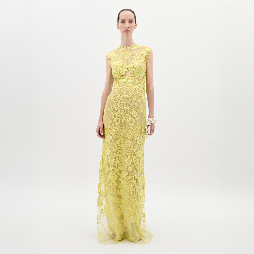 Una persona se encuentra de pie sobre un fondo blanco liso con el Vestido Aika, un vestido de encaje amarillo, largo hasta el suelo, sin mangas y con intrincados estampados. Lleva el cabello recogido y un brazalete floral blanco adorna su muñeca derecha.