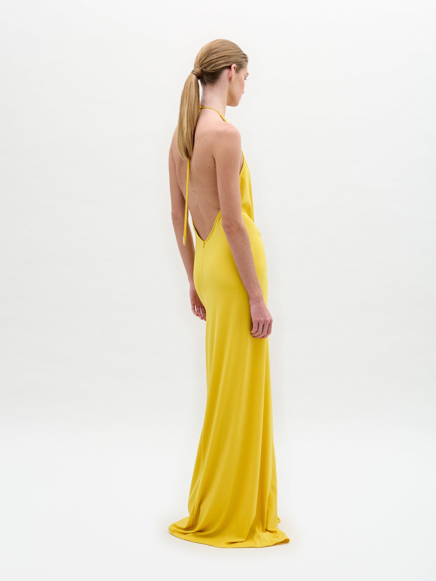 Una persona con el pelo largo y liso recogido en una coleta baja lleva el vestido amarillo sin mangas y sin espalda Aiko Dress, fabricado en Colombia, de pie y de lado sobre un fondo blanco liso.