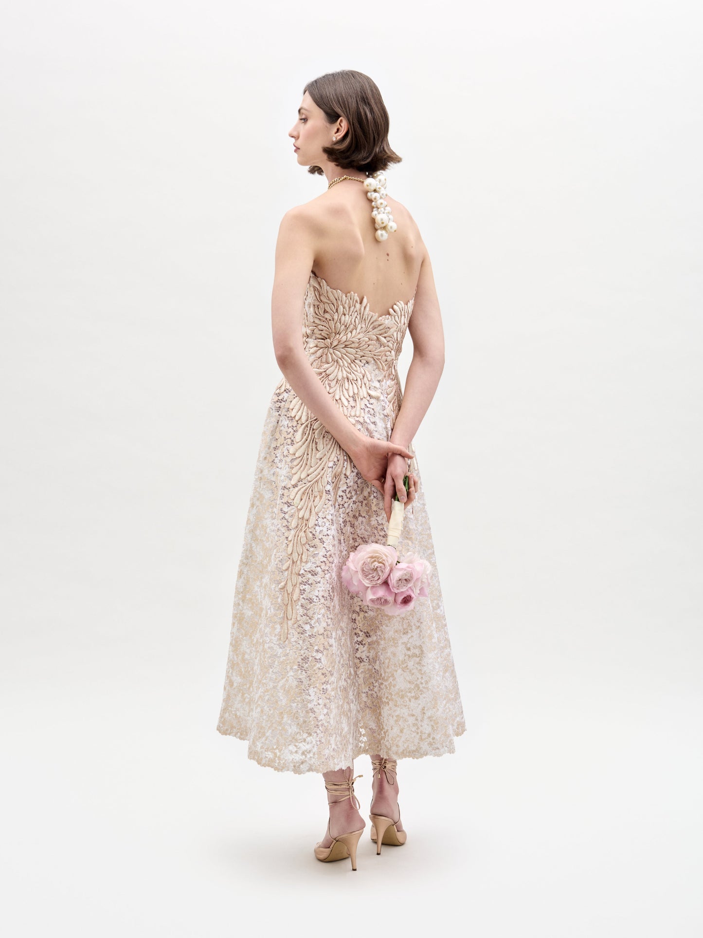 Una mujer con un vestido Alejandra beige bordado posa de espaldas a la cámara, sosteniendo un ramo de rosas rosadas. Lleva el pelo corto y tacones sobre un fondo blanco liso.