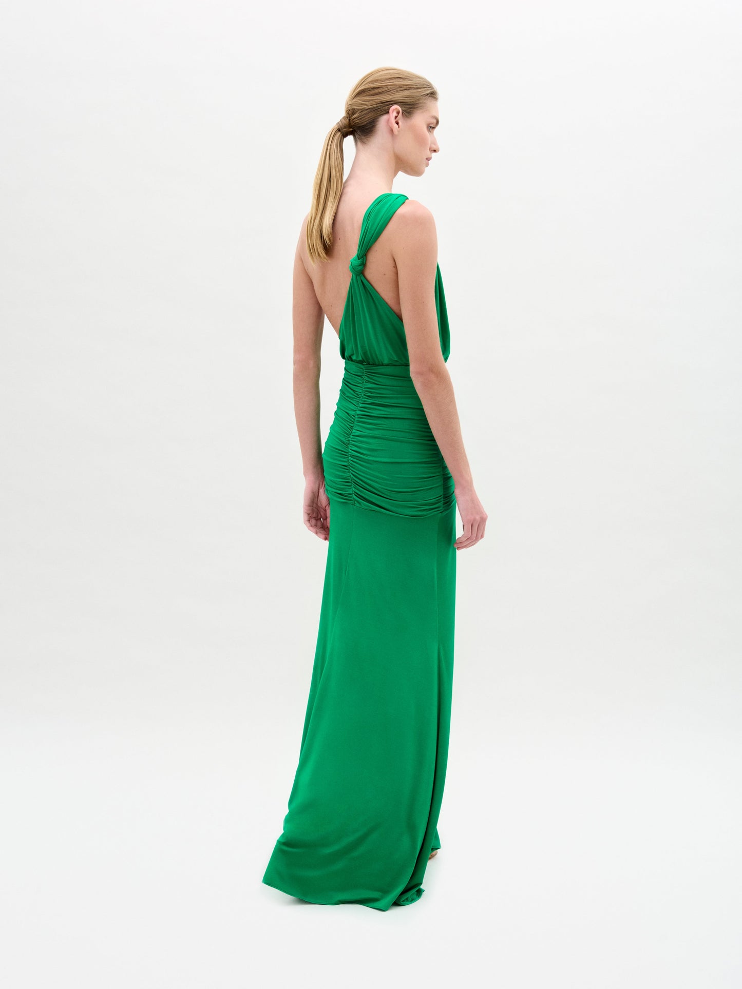 Una persona posa de espaldas a la cámara con el cabello recogido en una coleta baja, luciendo el Antara Dress: un vestido maxi verde con detalle fruncido en la espalda y escote asimétrico, sobre fondo blanco.