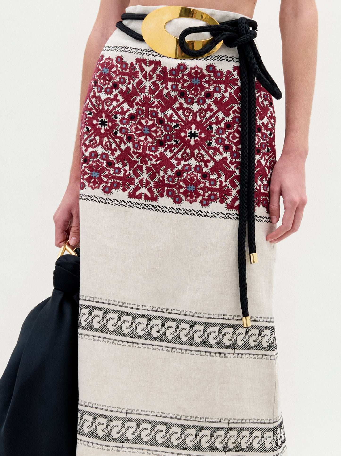 Una persona lleva la Falda Atira beige con bordados rojos y negros, un cinturón negro de tela con una gran hebilla dorada y un bolso negro con asa dorada. Sólo se muestran el torso y la falda.