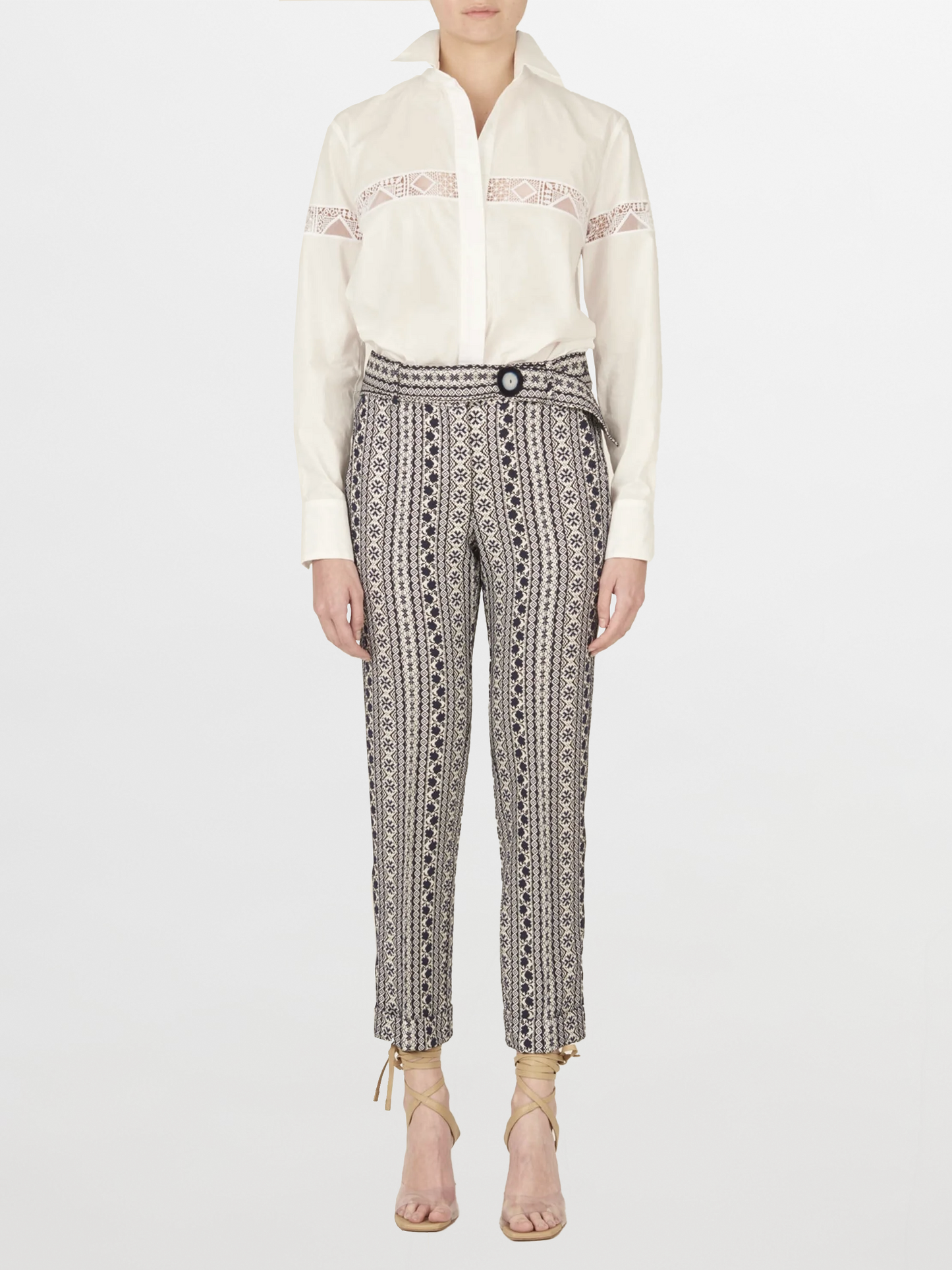 Una persona está de pie mirando hacia adelante con la Blusa Andrea White, que presenta un bordado intrincado, combinada con pantalones estampados de cintura alta y sandalias de tacón con cordones de color beige sobre un fondo blanco liso.