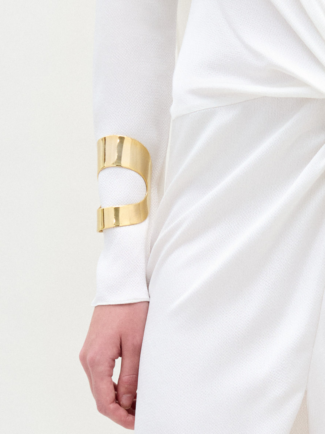 Persona vestida con una prenda blanca texturizada de manga larga con tejido fruncido en la cintura y el Cuff Artemis, una gran pulsera brillante baño de oro, en el antebrazo. Sólo son visibles la parte inferior del torso y un brazo.