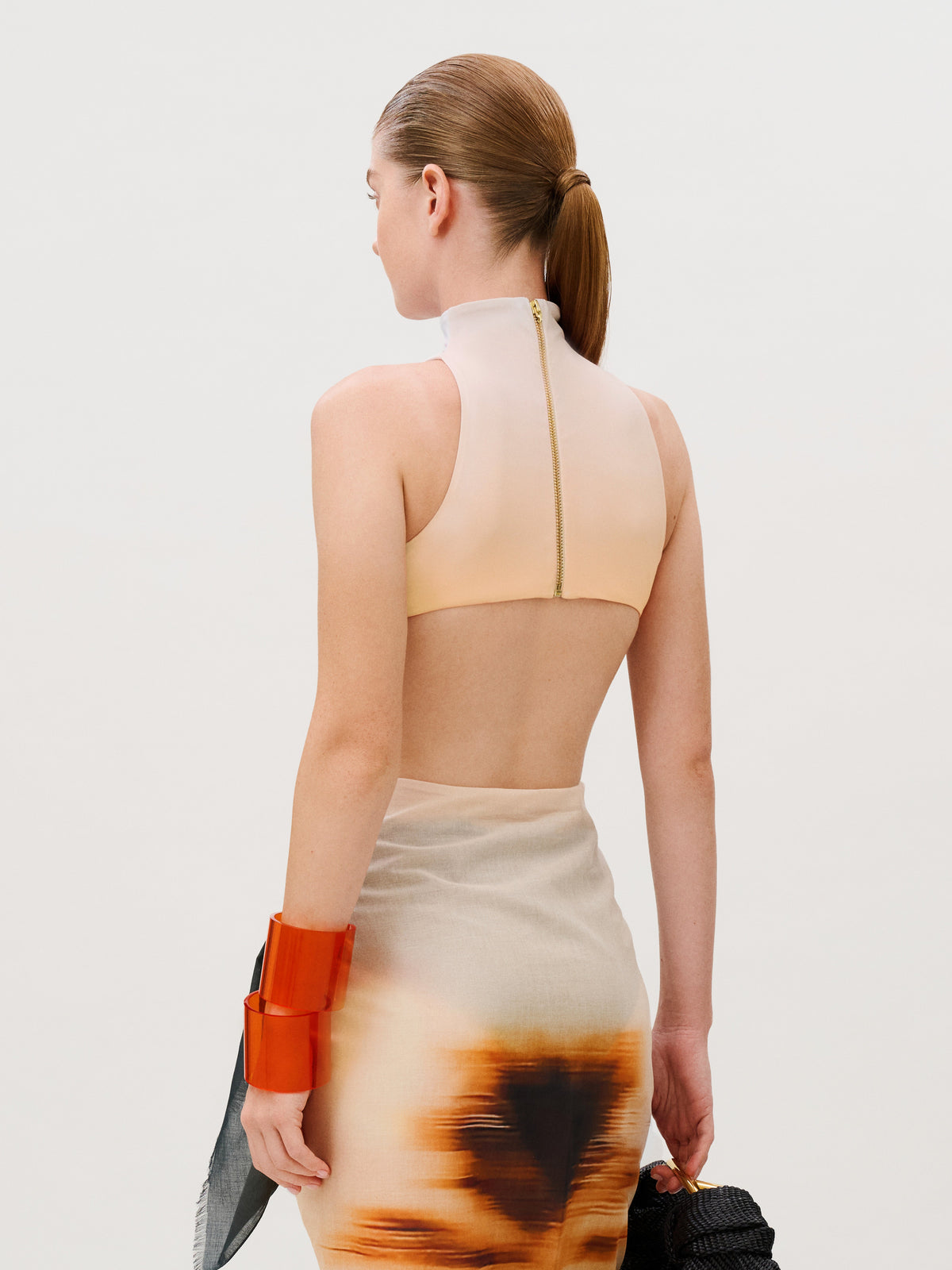 A model wears the Asia Swim Bodysuit, a high-neck Lycra bodysuit with a back zipper and a skirt in an abstract brown and beige print, accented by a bright orange cuff bracelet. Made in Colombia.