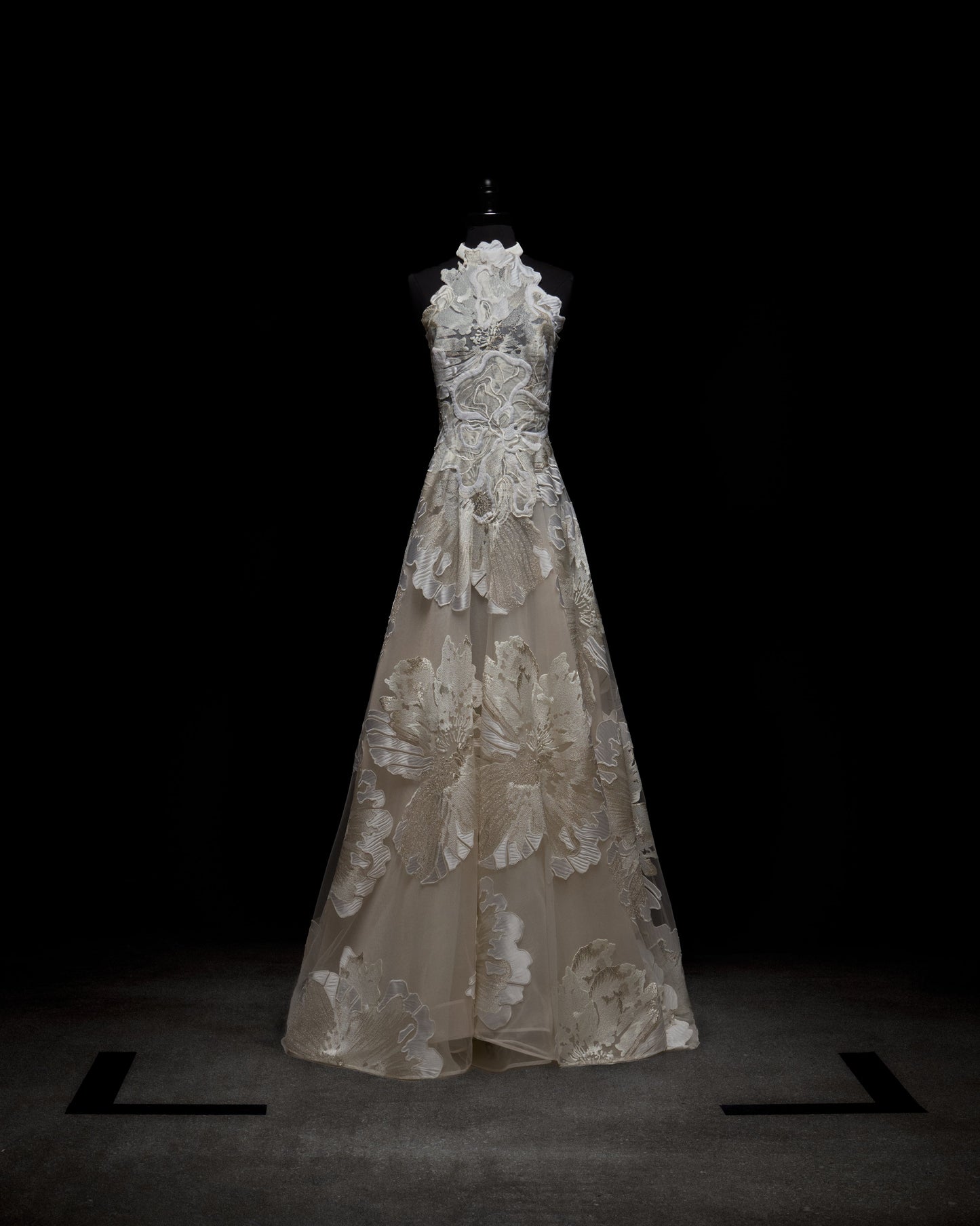 El vestido Becca, un vestido blanco sin mangas, exhibido sobre un maniquí negro contra un fondo oscuro, luce intrincados detalles de encaje y estampados florales. Este vestido presenta un escote alto con un corpiño ajustado que se extiende hasta una elegante falda hasta el suelo.