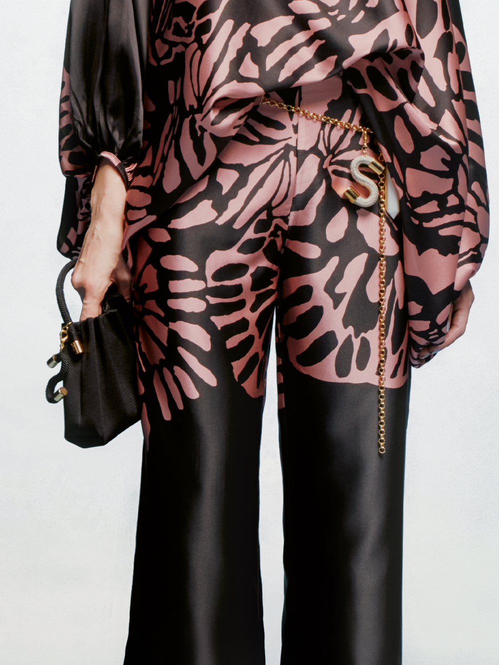 Persona con pantalón negro Andie y top holgado con un atrevido estampado abstracto rosa, sosteniendo un pequeño bolso negro y llevando un cinturón de cadena con un gran colgante dorado en forma de "S". La foto está recortada por el torso.