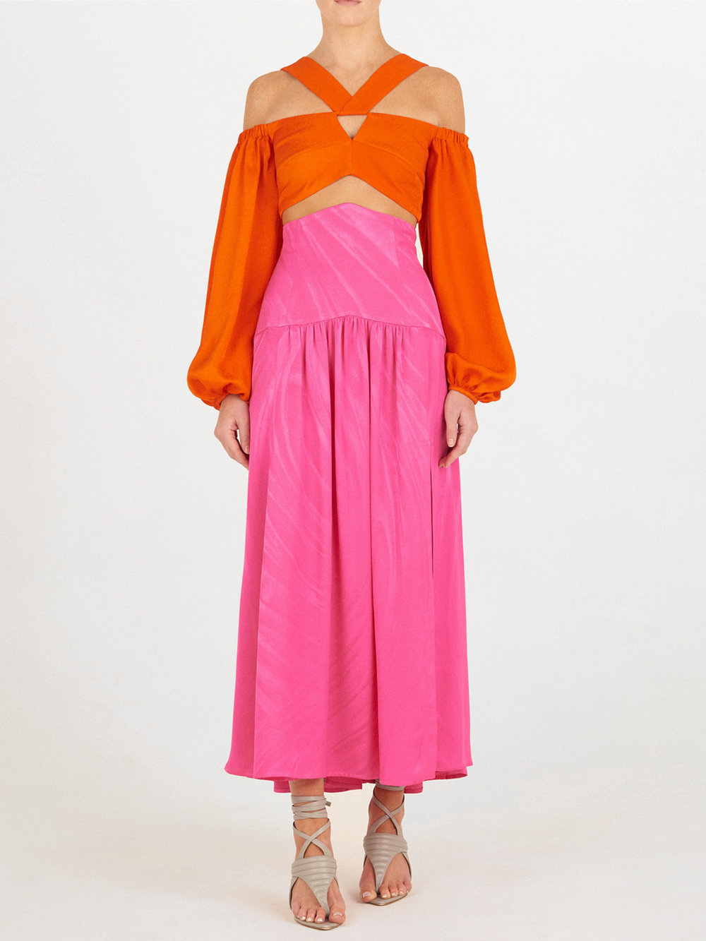 Una persona, sobre un fondo liso, lleva la Falda Callie Fuchsia, una falda de jacquard de alta costura hasta el suelo, combinada con un top naranja de hombros recortados, manga larga y tiras cruzadas, y sandalias de tiras beige.