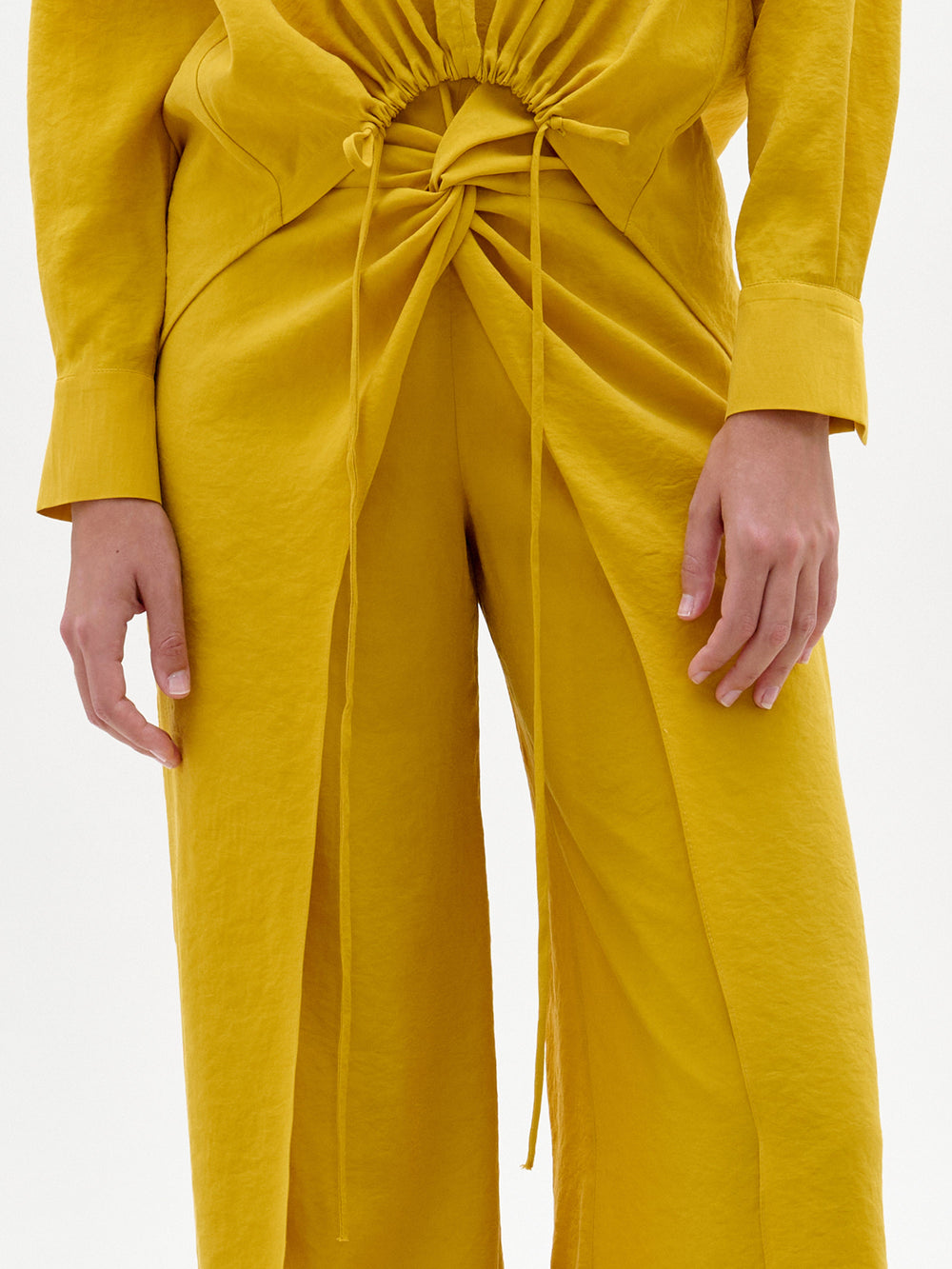 Una persona lleva el pantalón Canturipe, de talle alto, color amarillo mostaza, con cintura fruncida y lazos delanteros, combinado con la camiseta de manga larga a juego. Sólo se ven el torso y las manos. Este elegante conjunto está fabricado en Colombia.