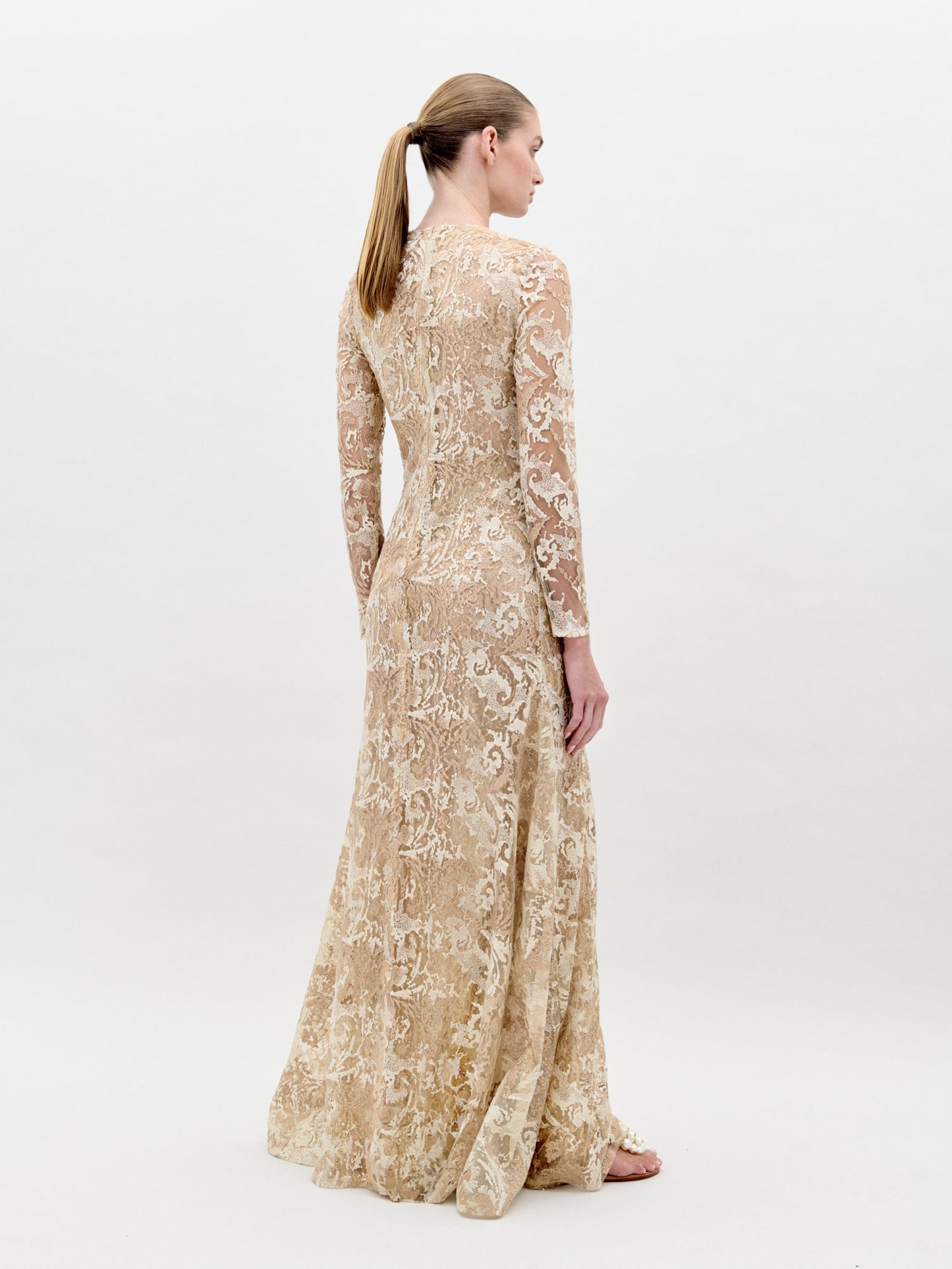 A woman in the Nila Dress Gold, a beige, floor-length, long-sleeved lace gown with a keyhole neckline, stands against a plain background, ready for pre-order ahead of the ship-by date on February 1st, 2025.