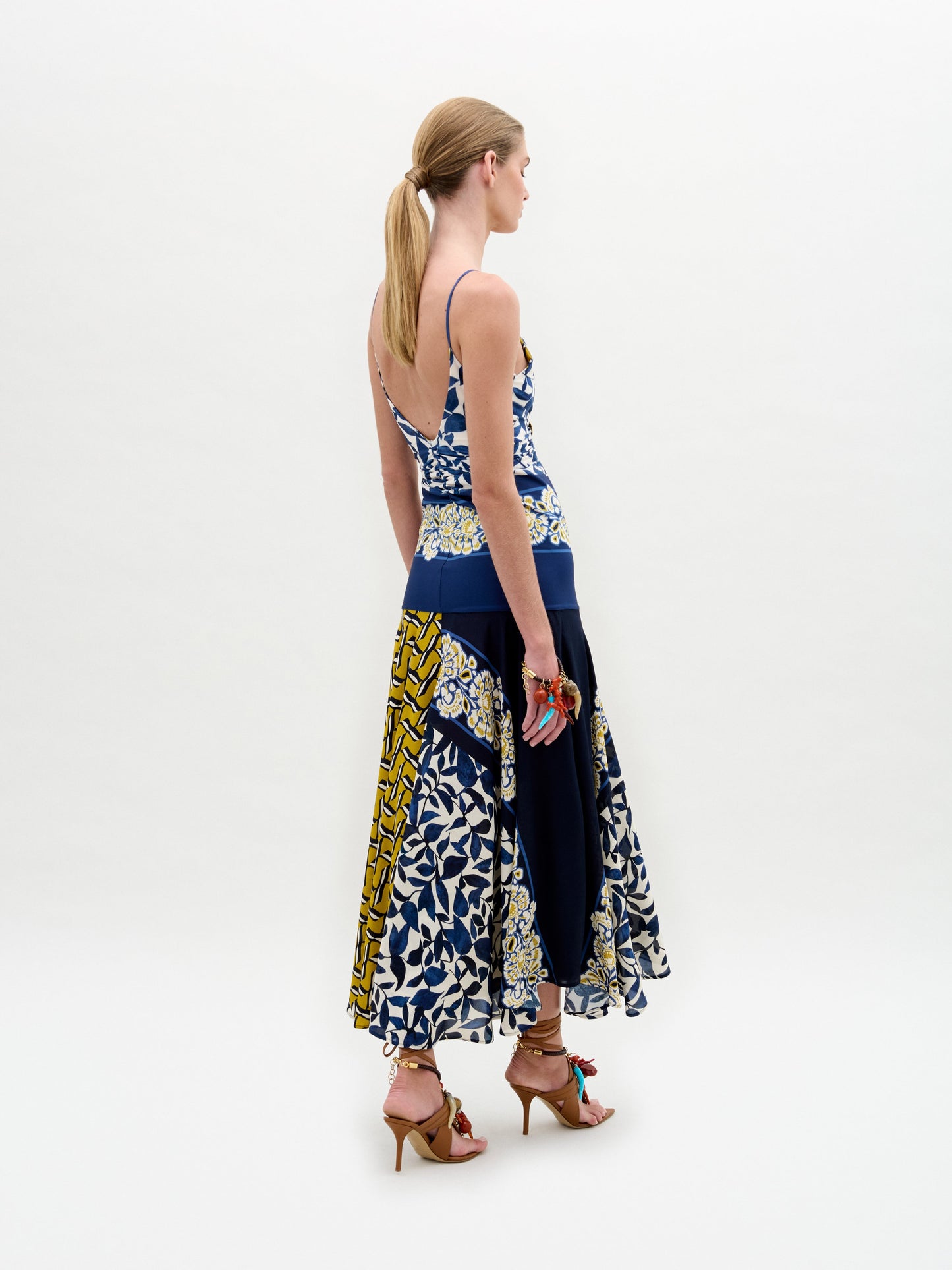 Una mujer con coleta baja y sandalias marrones de tacón alto luce el vestido Casiana, un midi entallado con tirantes finos, falda acampanada y estampado azul, blanco y amarillo. Está de espaldas sobre un fondo blanco liso.