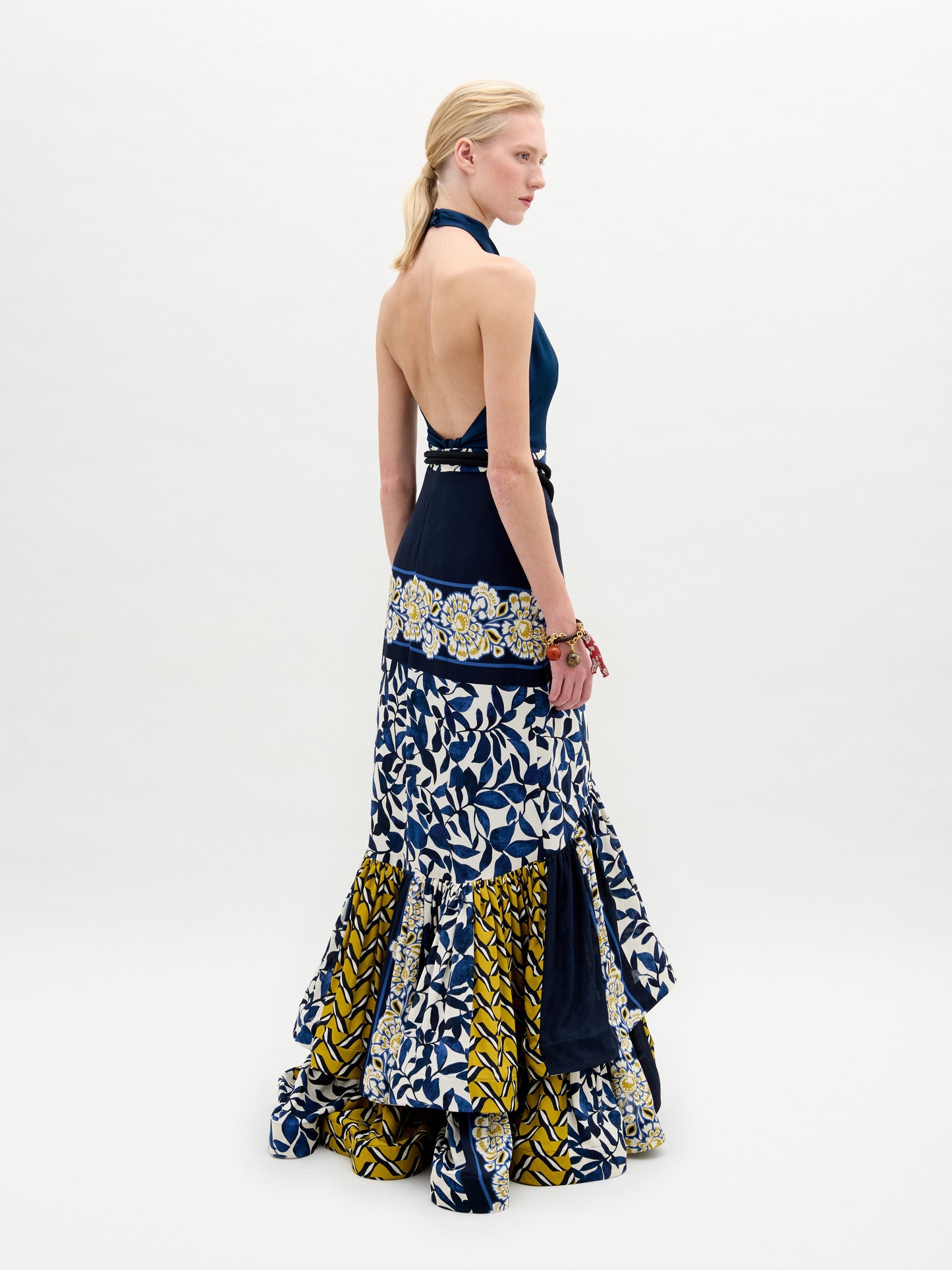 Una mujer de piel clara y pelo rubio sobre un fondo liso luce la falda Elliot, una maxifalda sin espalda y cuello halter con motivos geométricos y florales en azul, blanco y amarillo y diseño escalonado con volantes.