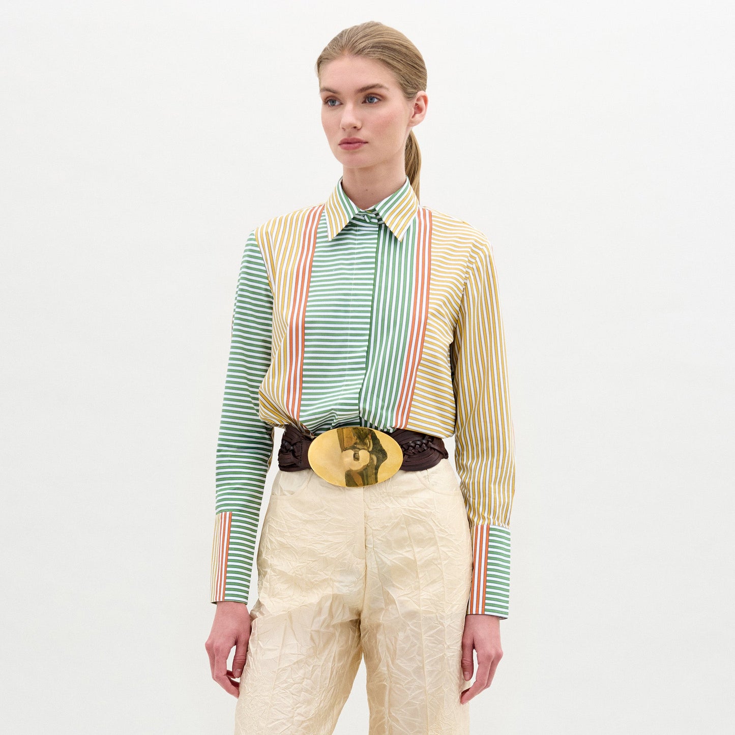Una mujer mira hacia adelante sobre un fondo liso, vistiendo la Camisa Cubillos (100% algodón, a rayas), pantalones color crema y un gran cinturón marrón con hebilla ovalada dorada. Tiene una expresión neutra.