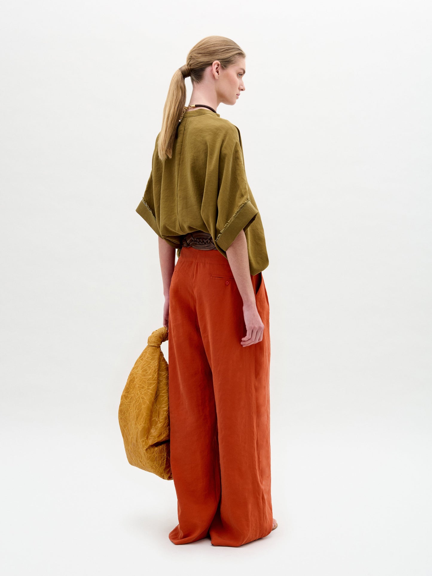Una persona de espaldas, vestida con la blusa Daia verde oliva y pantalones anchos naranja óxido, sostiene un gran bolso amarillo con textura. Lleva el pelo rubio recogido en una coleta baja sobre un fondo blanco liso.