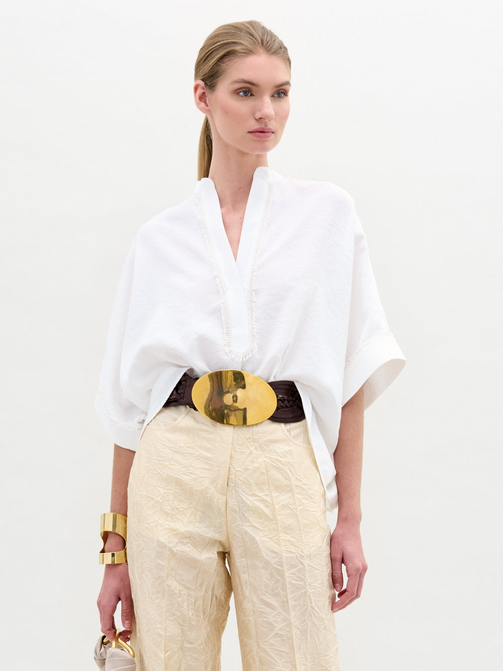 Una mujer con una blusa Daia y pantalones color crema se encuentra de pie frente a un fondo sencillo. Lleva un cinturón con una gran hebilla dorada, sostiene un bolso y luce brazaletes dorados gruesos, mostrando un estilo elegante que parece hecho en Colombia.