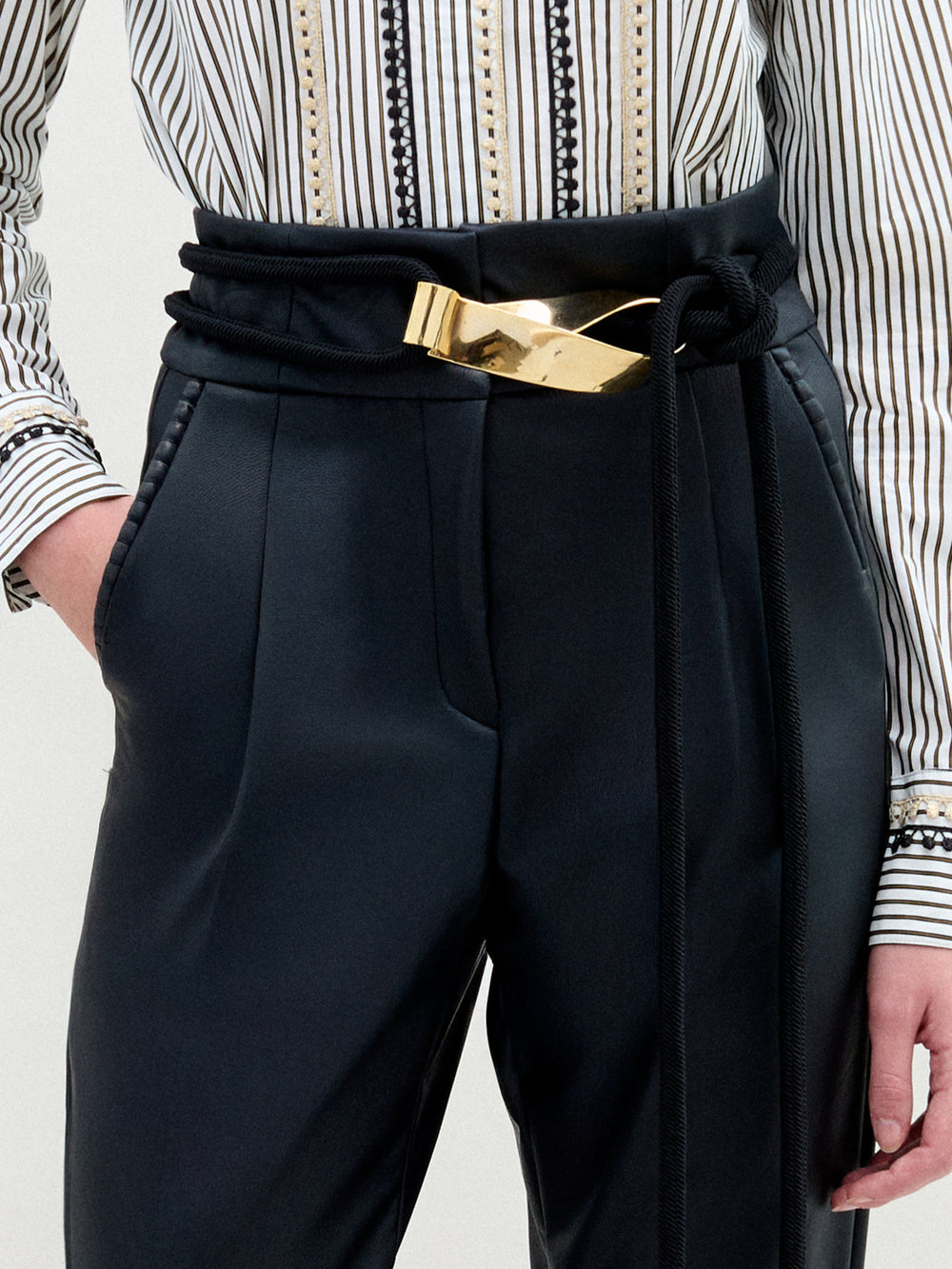Una persona combina unos pantalones negros de talle alto con el cinturón Daiana, con diseño de cordón y hebilla dorada, y una camisa de rayas de manga larga con puños decorativos y detalles delanteros. Una mano se introduce despreocupadamente en un bolsillo.