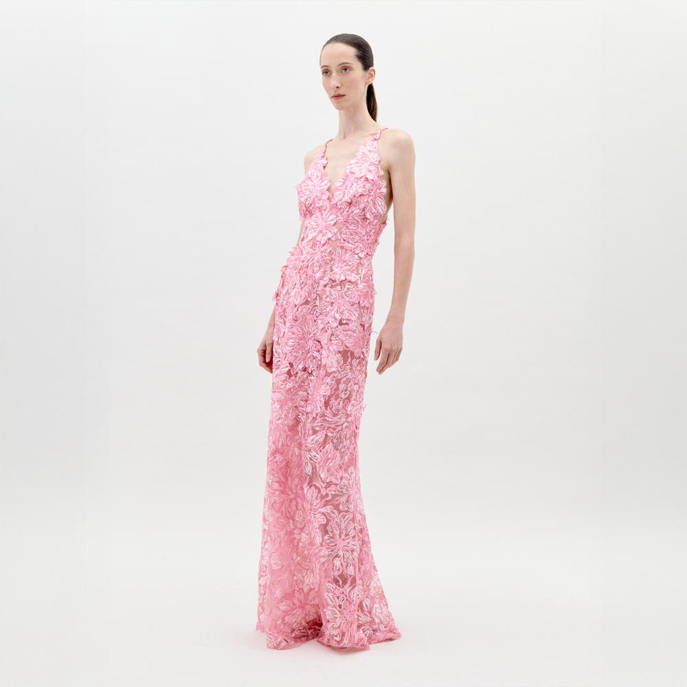 Una persona se encuentra de pie sobre un fondo claro y liso con un vestido Daira rosa hasta el suelo, con detalles florales texturizados y un delicado tul de encaje. Lleva el cabello largo y liso recogido hacia atrás y su expresión es neutra.